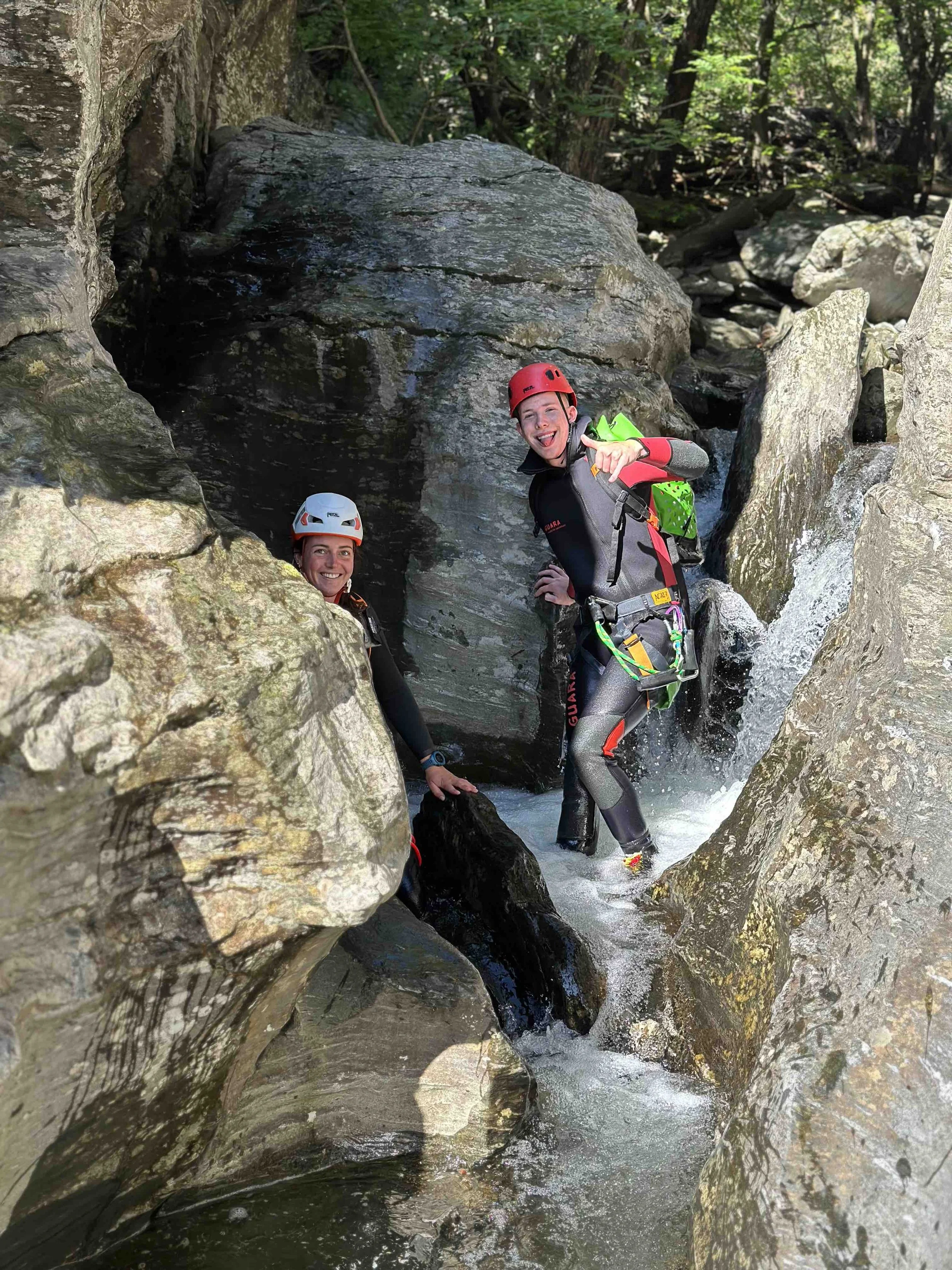 Deux personnes en équipement de spéléologie dans une rivière de montagne, portant des casques rouges et noirs, utilisant des combinaisons résistantes à l'eau, au milieu de rochers et cascade d'eau, souriant et posant pour la photo.