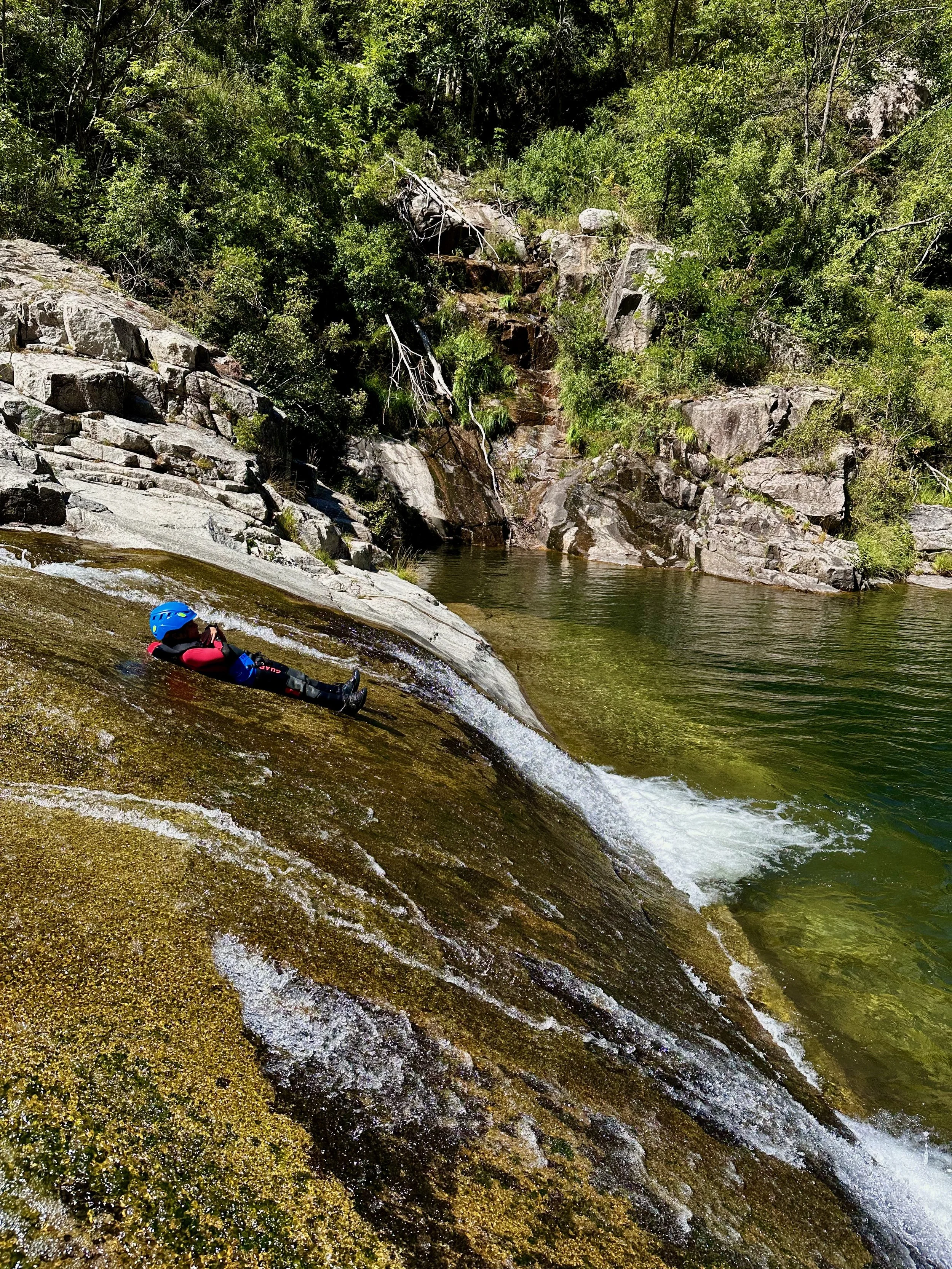 Personne faisant du canyoning sur une roche glissante dans une rivière entourée de végétation et de rochers.