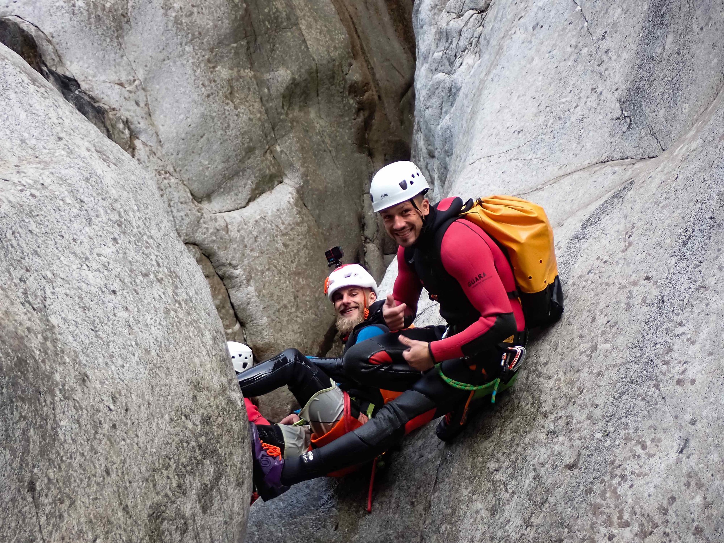 Deux hommes en équipement d'escalade dans une fissure rocheuse, souriants, donnant un signe de pouces, avec des casques de sécurité et des sacs à dos, dans un canyon ou une falaise.