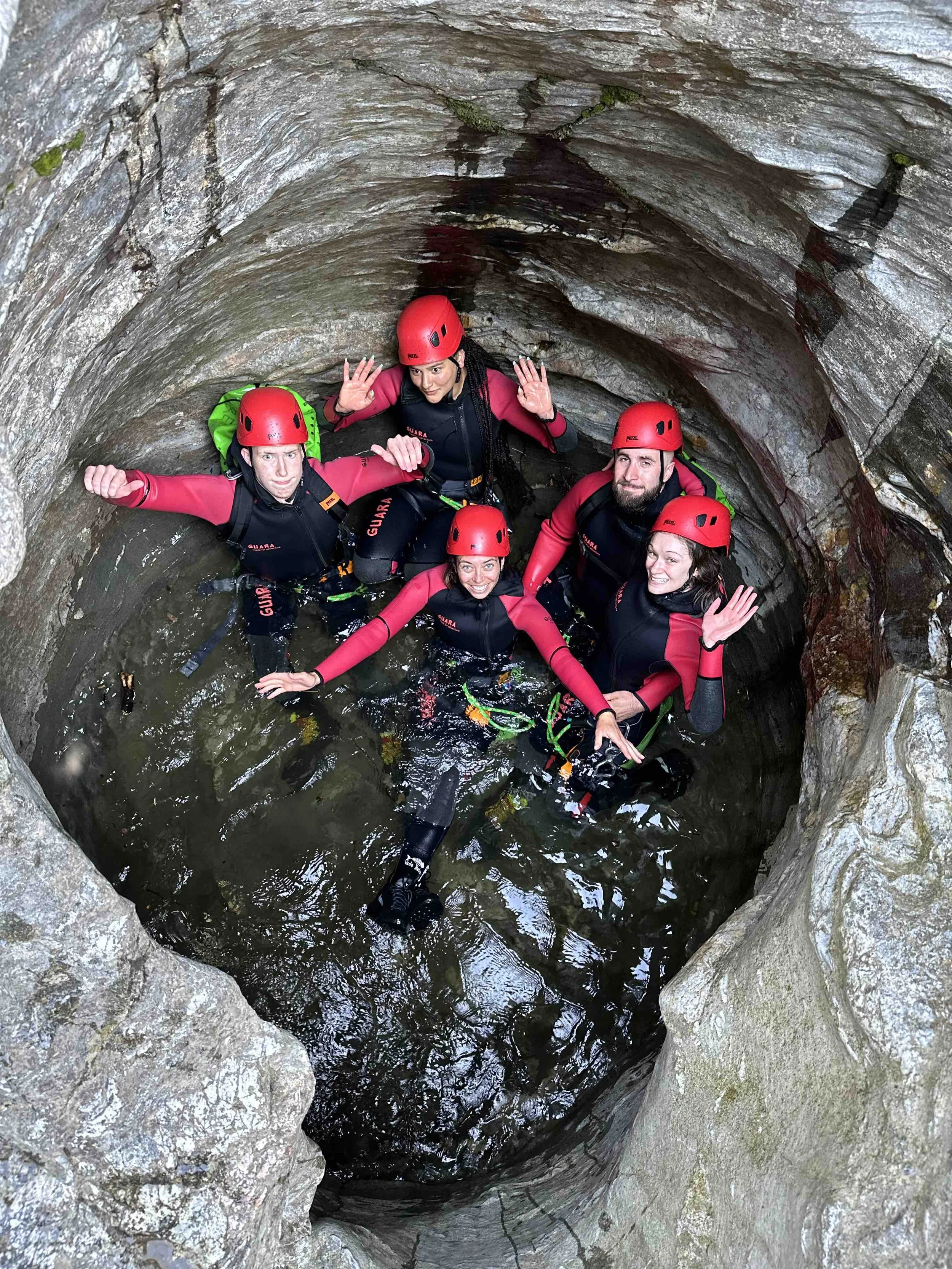 Cinq personnes équipées de casques rouges et de combinaisons noires et roses, en train de faire de la canyoning dans une gorge rocheuse, en se tenant dans l'eau.