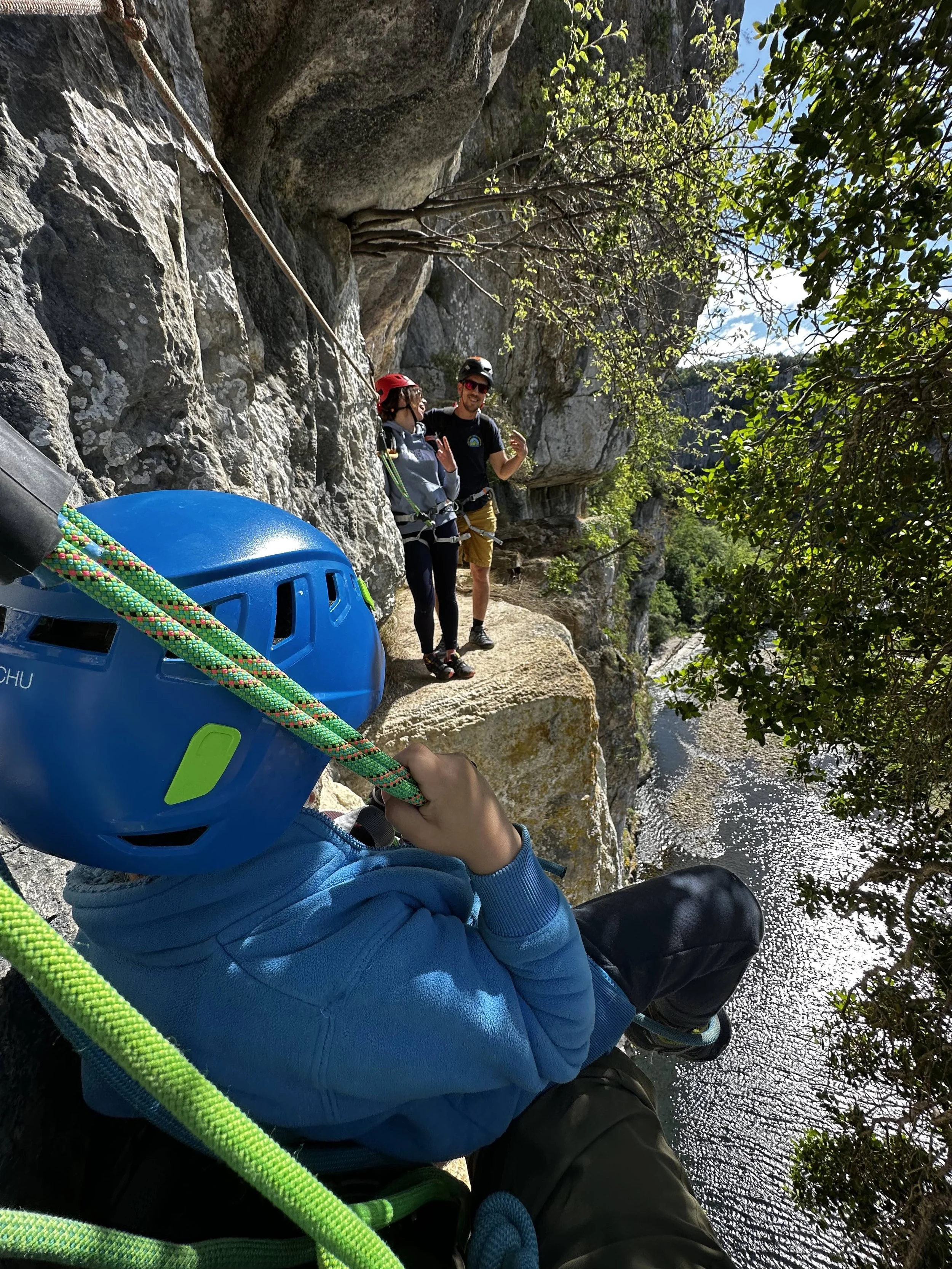 Une personne portant un casque bleu et des vêtements bleus tient une corde de escalade lors d'une excursion en canyoning ou via ferrata le long d'une falaise rocheuse près d'une rivière, avec deux autres personnes en arrière-plan qui parlent et portent des équipements de sécurité.