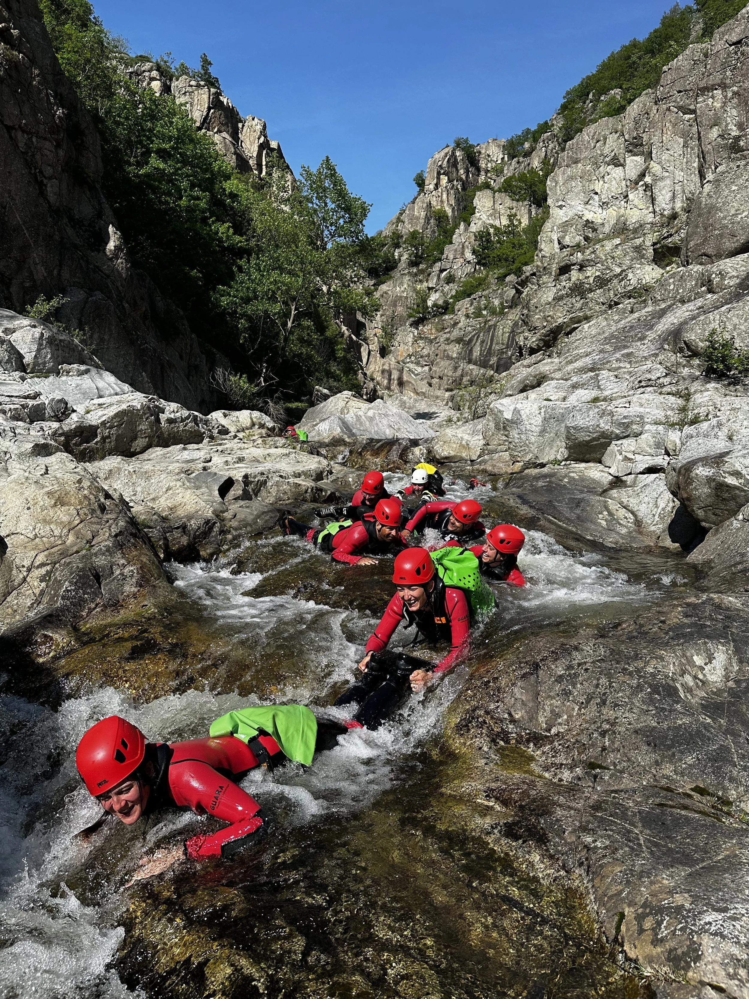 Groupe de personnes en équipements de slalom avec casques rouges et vestes rouges, faisant du canyoning dans une rivière rocheuse en montagne, entourée de falaises et de végétation, sous un ciel bleu.