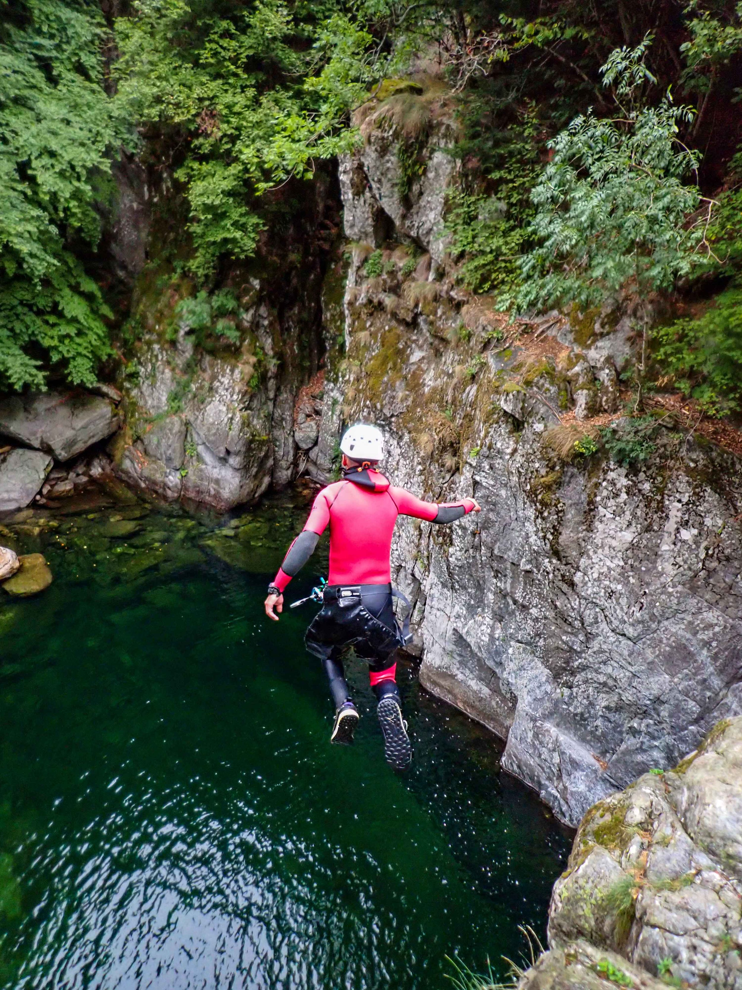 Une personne en tenue de canyoning avec casque, sautant dans une rivière entourée de rochers et de végétation dense.