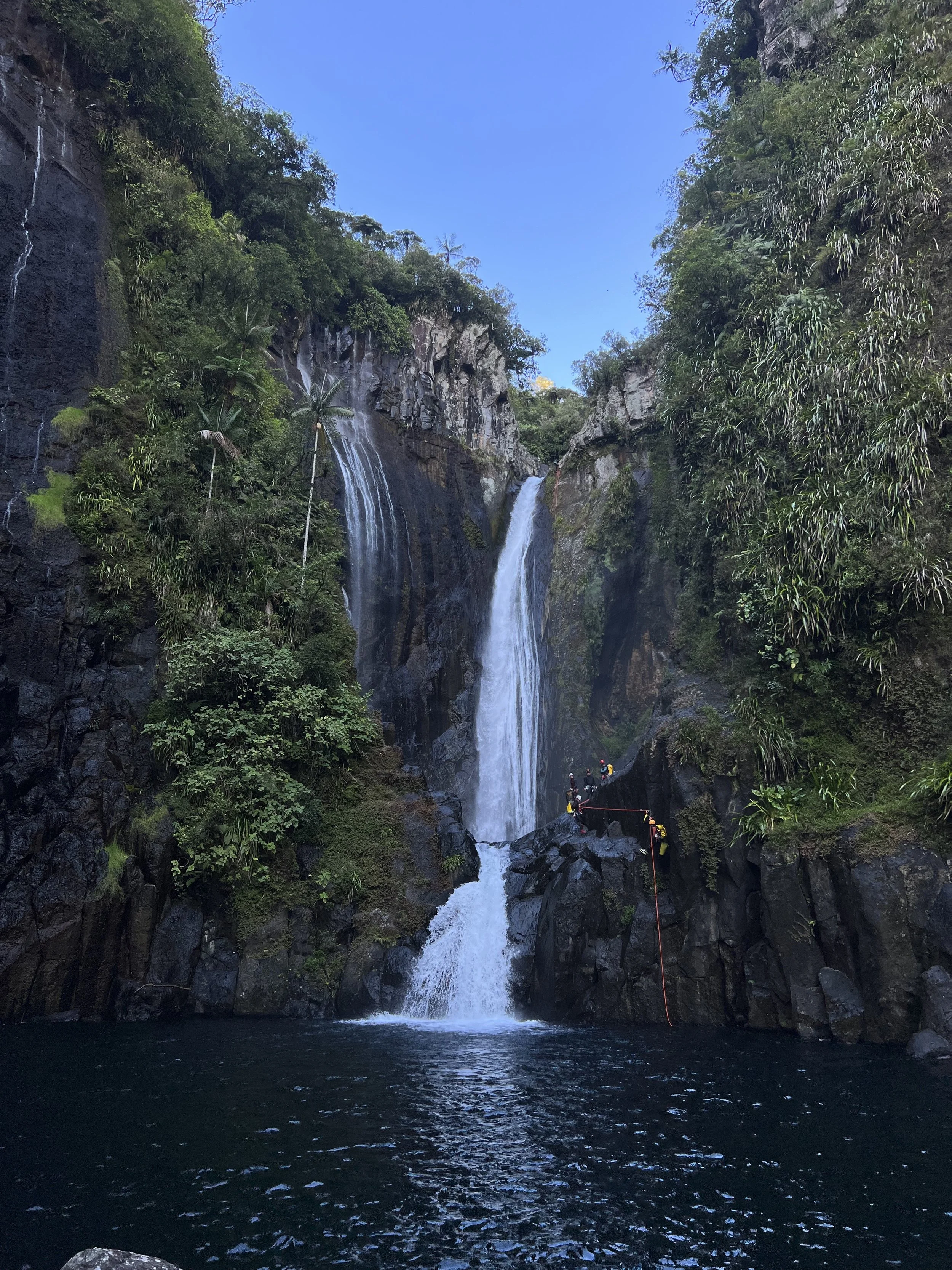 Cascade de plusieurs chutes d'eau dans une gorge rocheuse entourée de végétation dense sous un ciel bleu.