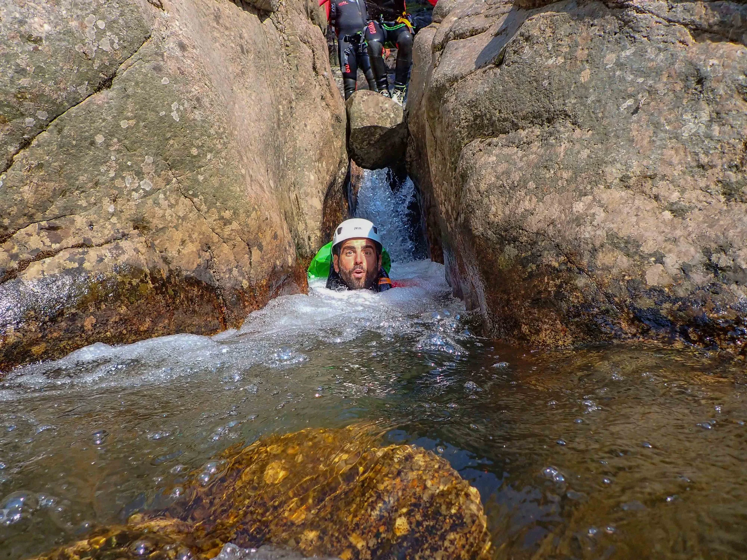 Un homme portant un casque blanc fait du canyoning dans une falie rocheuse, avec des groupes de personnes en arrière-plan.