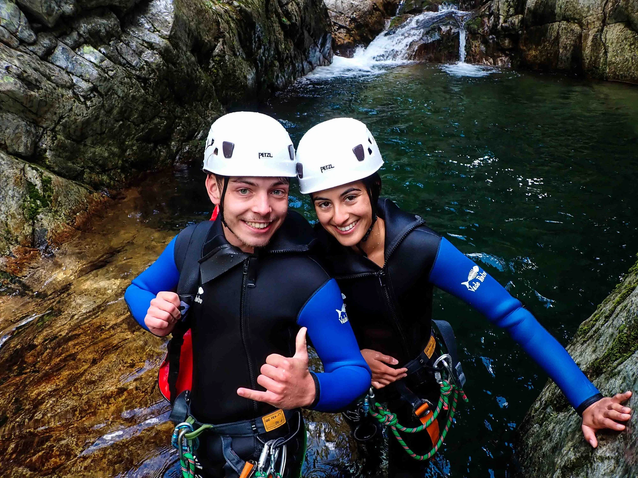 Deux personnes en équipement de canyoning avec casque et combinaisons vivent une aventure dans un gouffre, près d'une cascade et d'une rivière rocheuse.