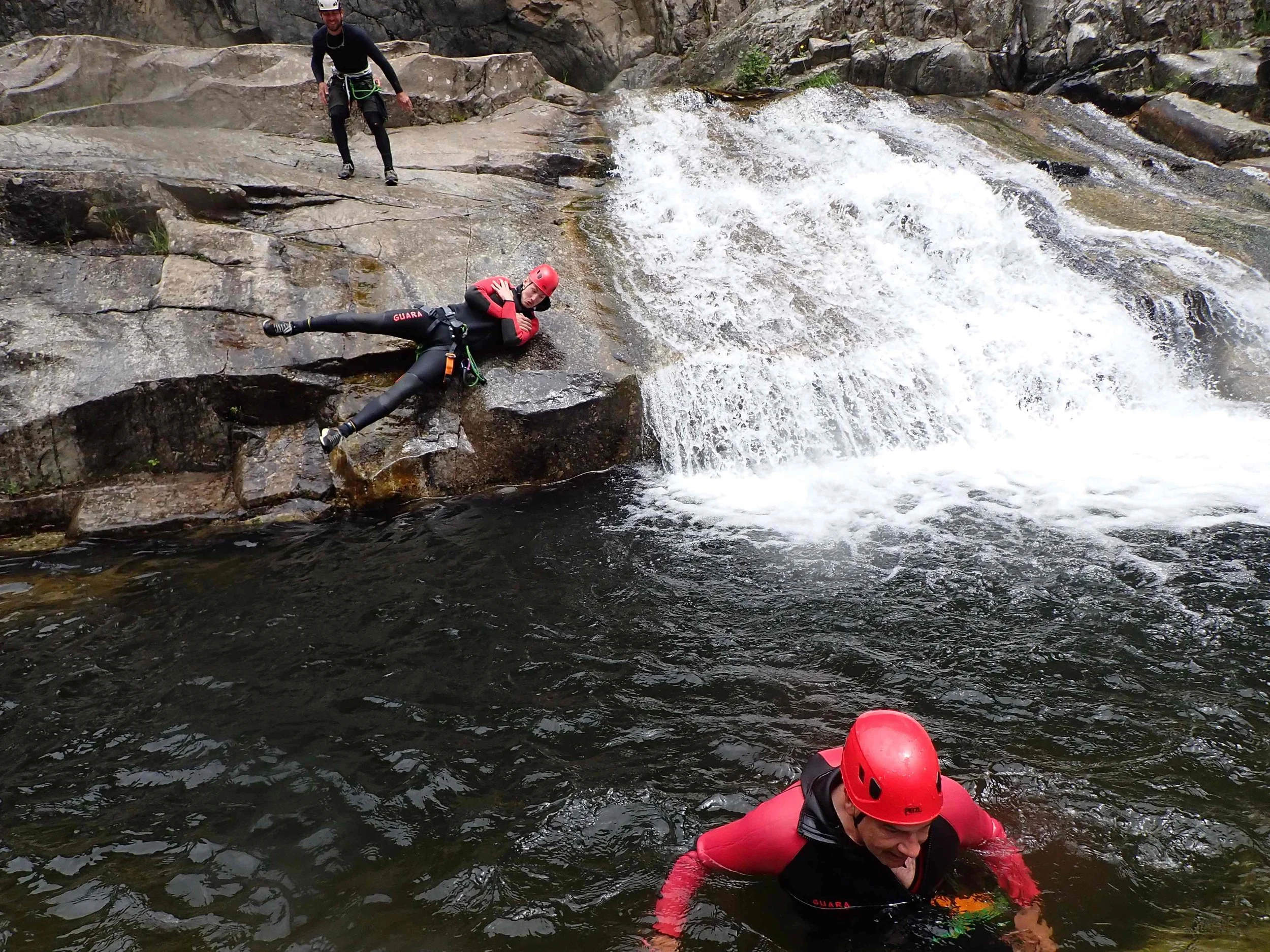 Trois personnes en équipement de sécurité, dont deux portant des casques rouges, en train de faire de la descente en eau vive sur une rivière rocheuse.