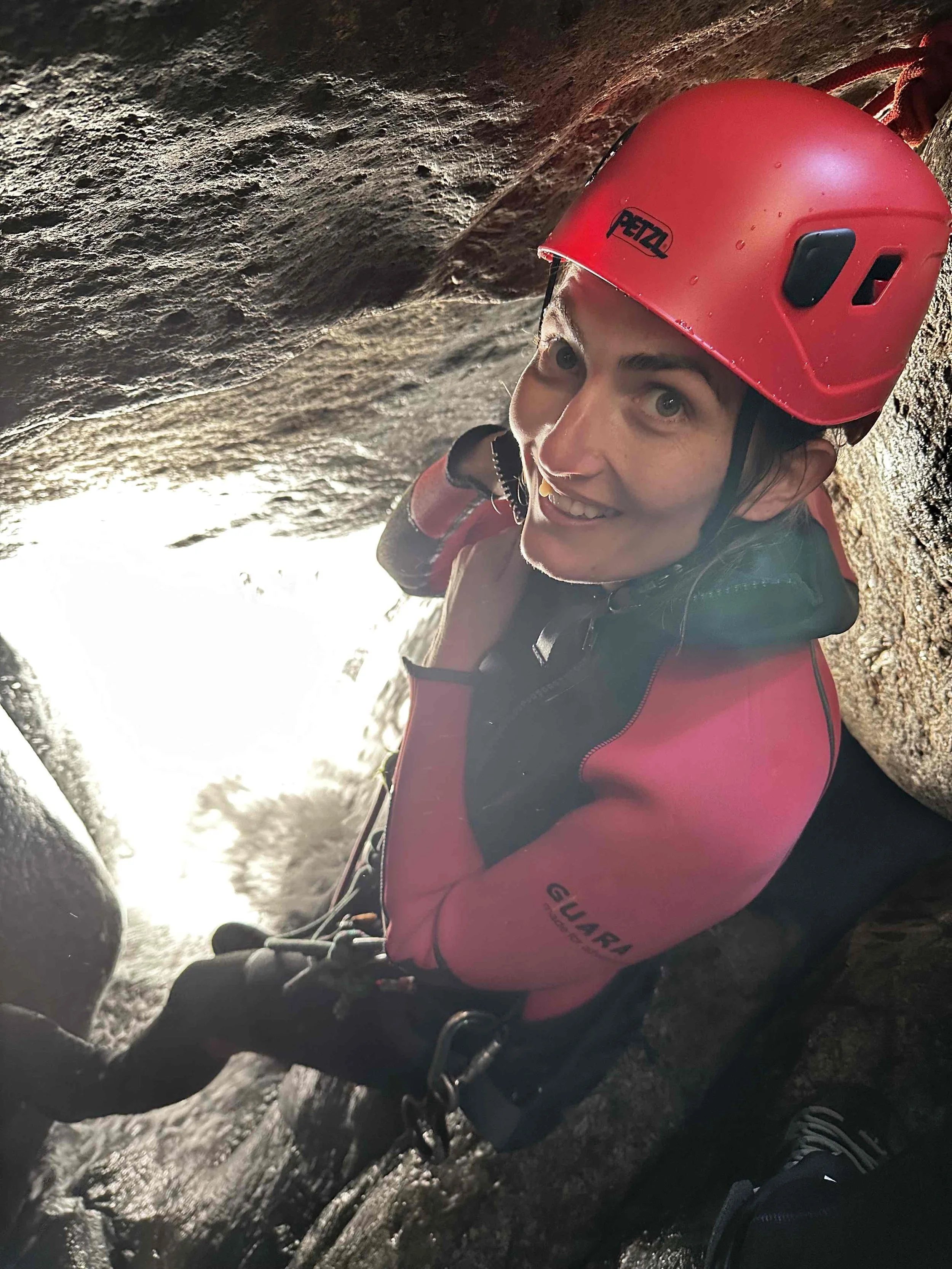 Une femme portant un casque rouge et un équipement d'escalade dans une grotte, souriant à la caméra.