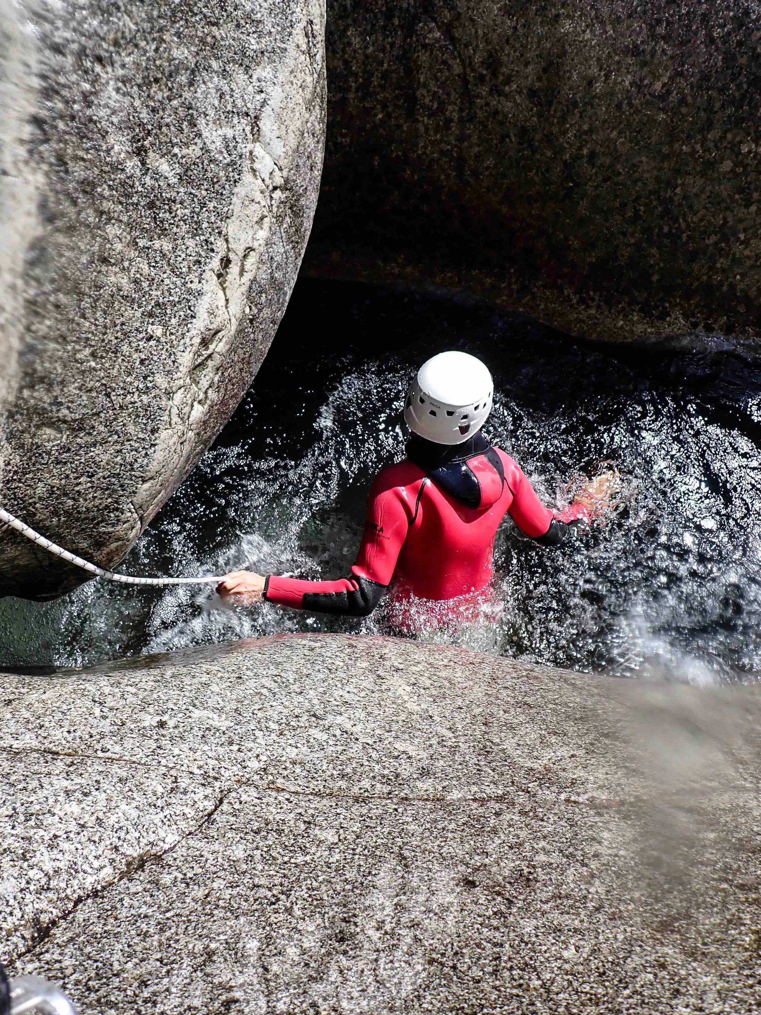 Personne portant un casque blanc et une combinaison de canyoning rouge, nageant dans une rivière entre des rochers.