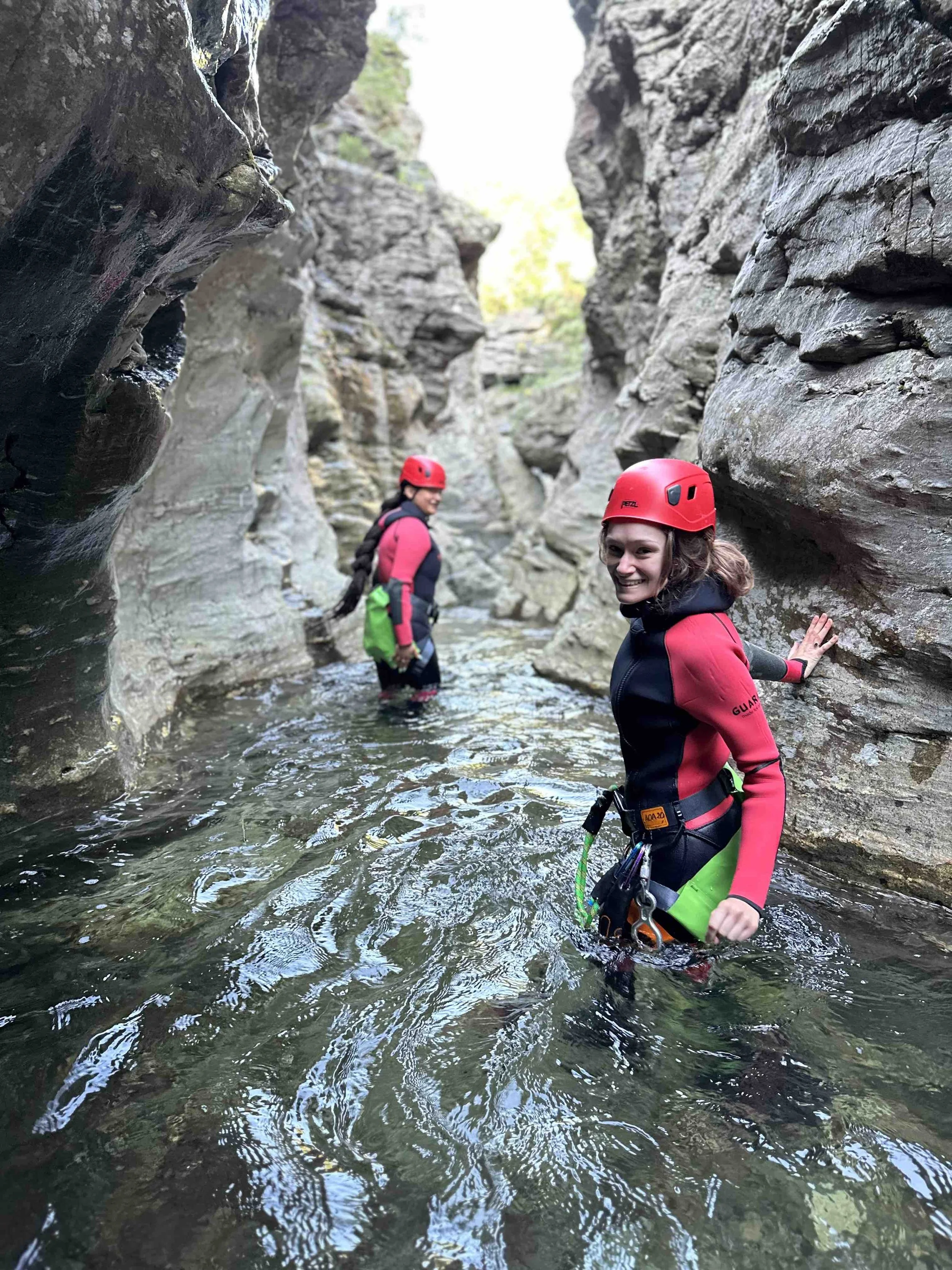 Deux femmes portant des casques rouges et des vêtements de randonnée sont debout dans une rivière entourée de falaises rocheuses.