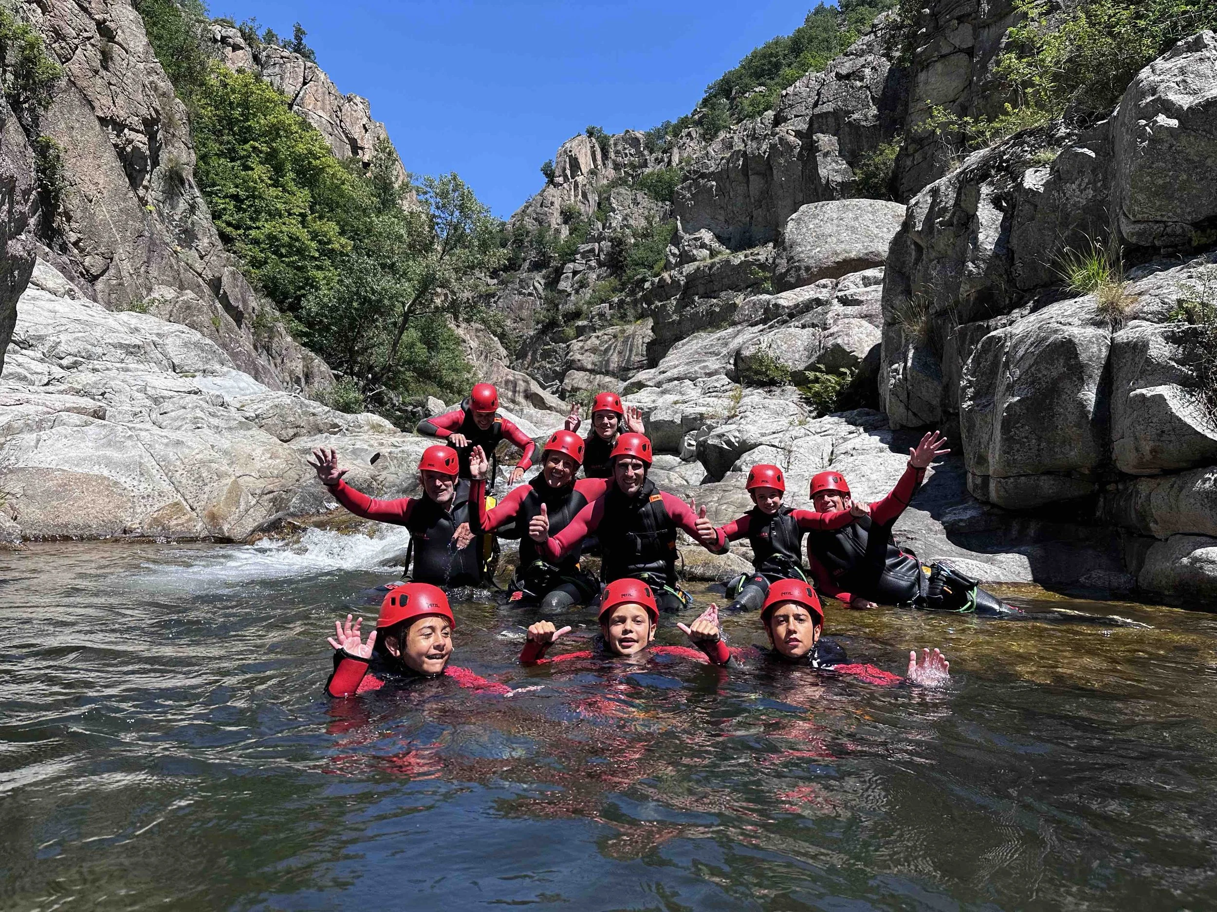 Groupe de personnes en équipement de canyoning avec casques rouges, dans une rivière entourée de rochers et de végétation, sous un ciel bleu.