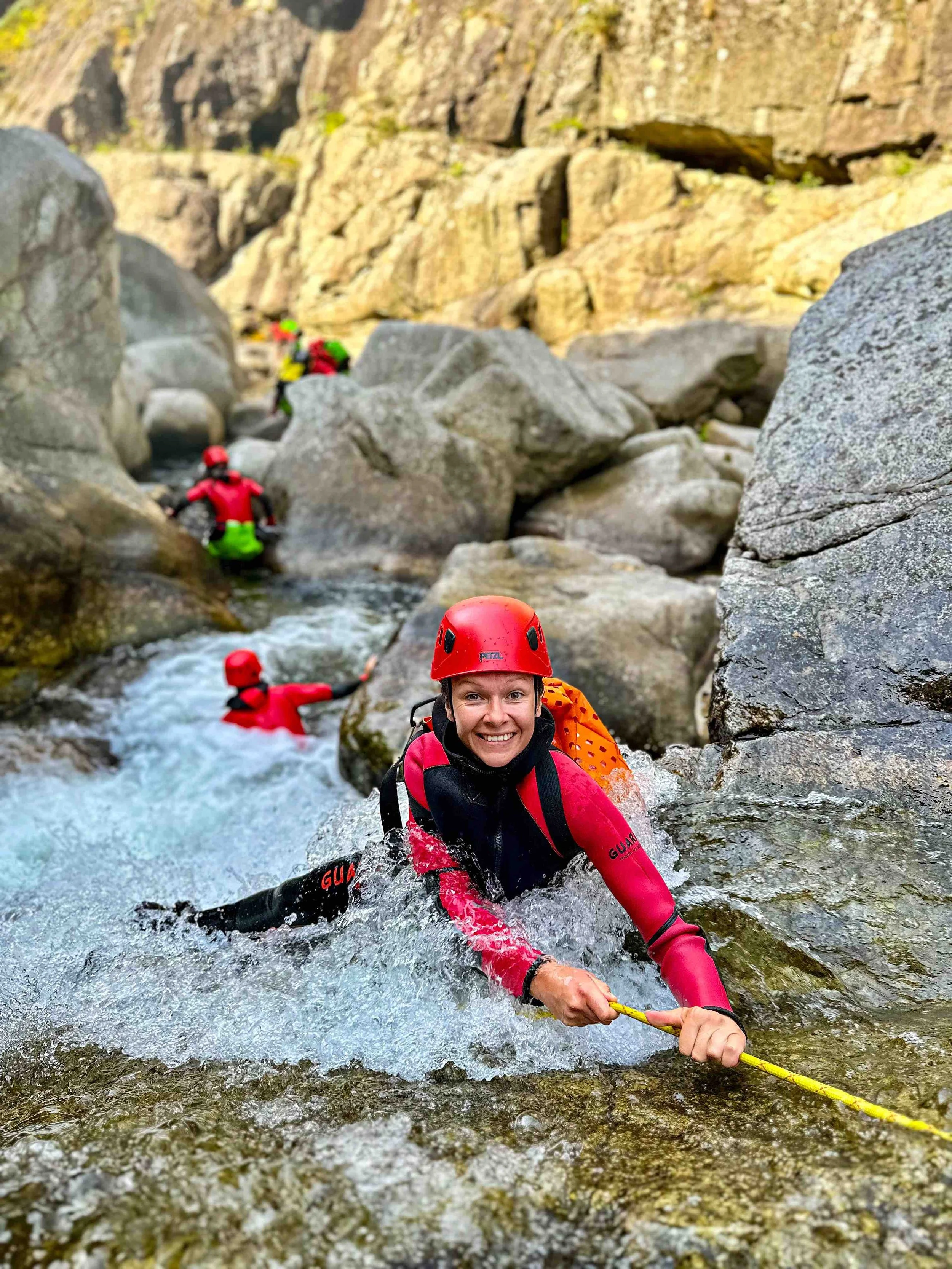 Une femme en équipement de canyoning, avec casque rouge et combinaison de protection, souriante en grimper sur des rochers dans une rivière de montagne, avec d'autres personnes en arrière-plan