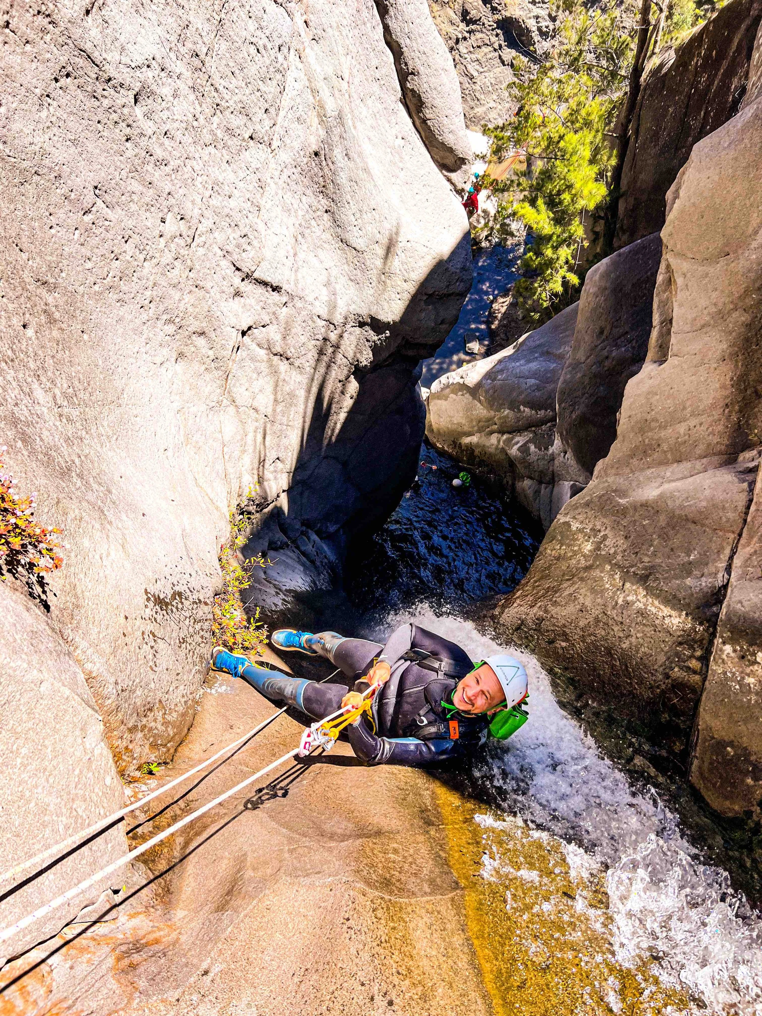 Personne faisant de l'escalade canyon dans une gorge rocheuse, portant un casque et une tenue de grimpe, tenant des cordes, avec des rochers et de l'eau en fond.