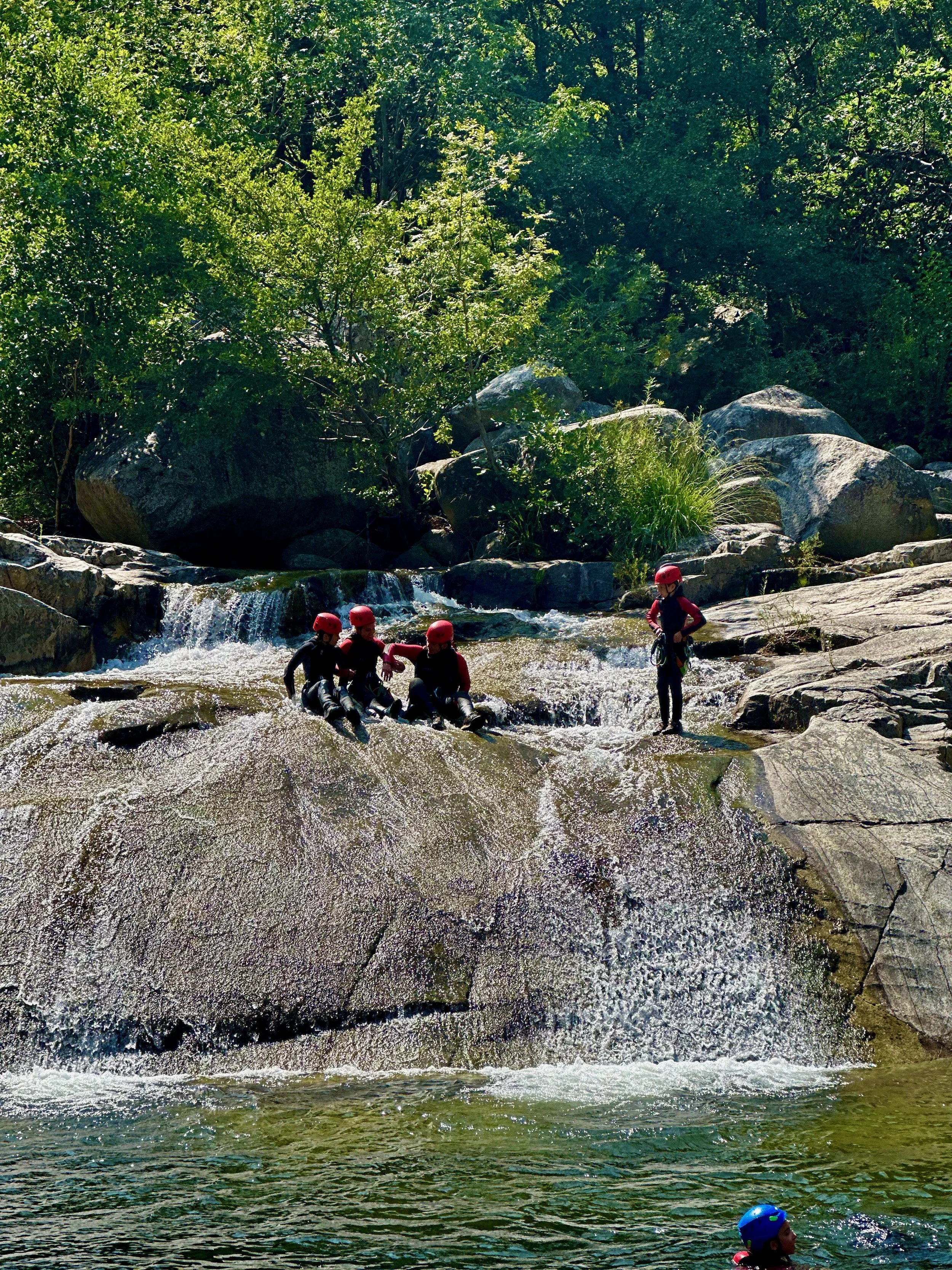 Quatre personnes en wetsuits et casques rouges près d'une cascade dans une rivière, entourées de verdure.