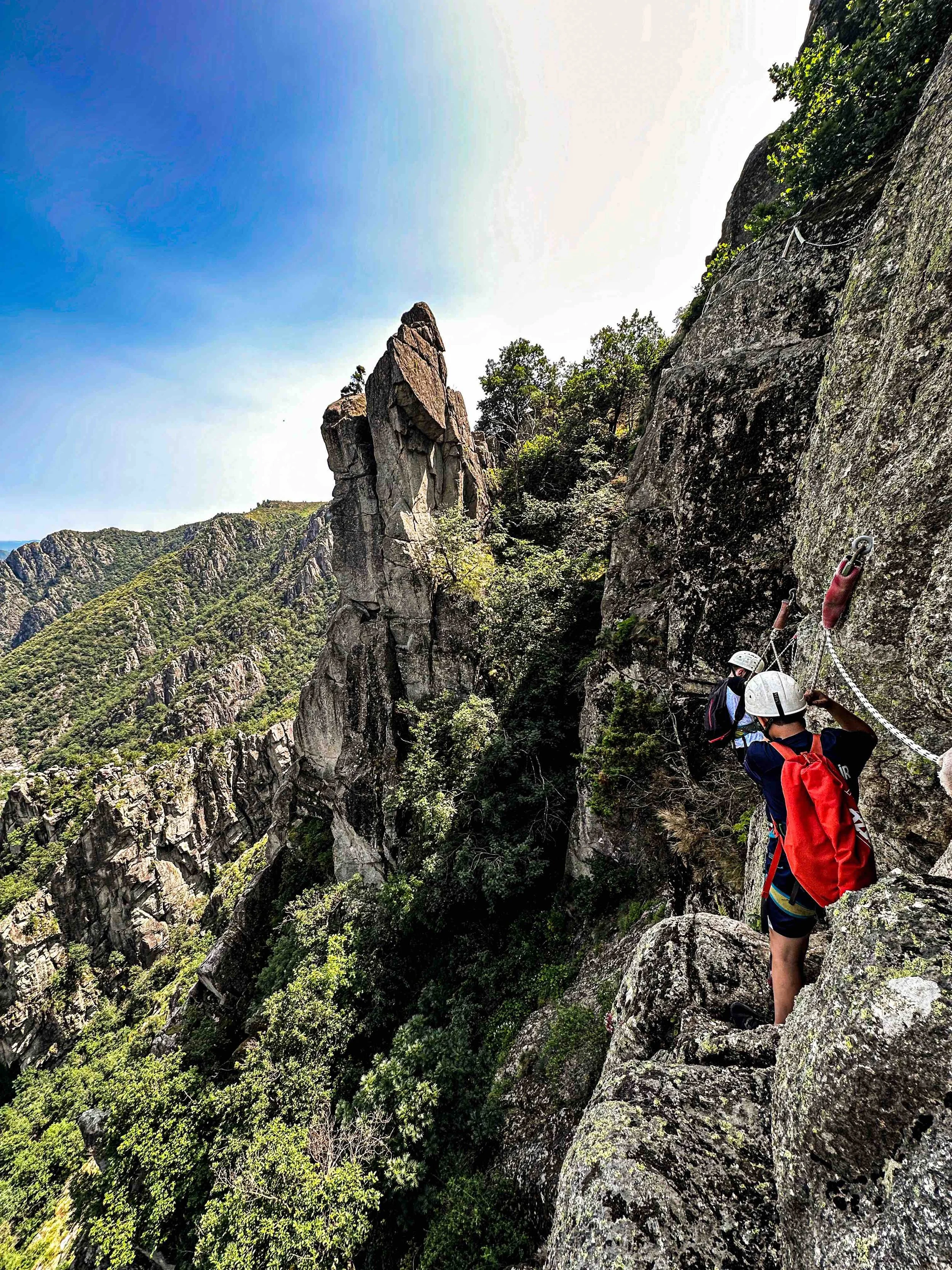 Escaladeurs portant des casques et un sac à dos en escaladant une falaise rocheuse avec vue sur une montagne verdoyante et un ciel bleu.
