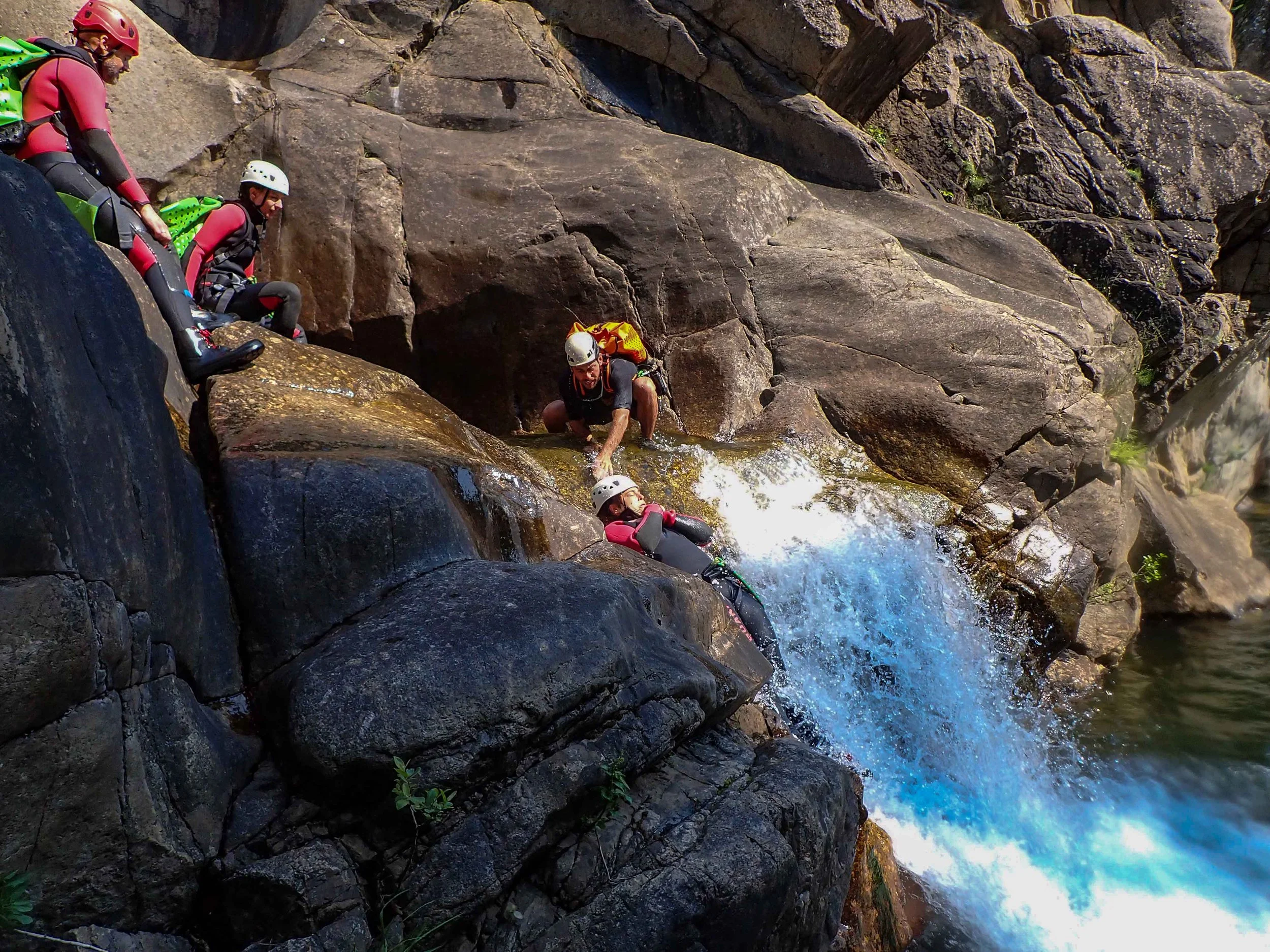 Des personnes faisant de la randonnée aquatique ou du canyoning, équipées de casques et de gilets de sauvetage, escaladant une roche près d'une cascade ou d'une rivière.