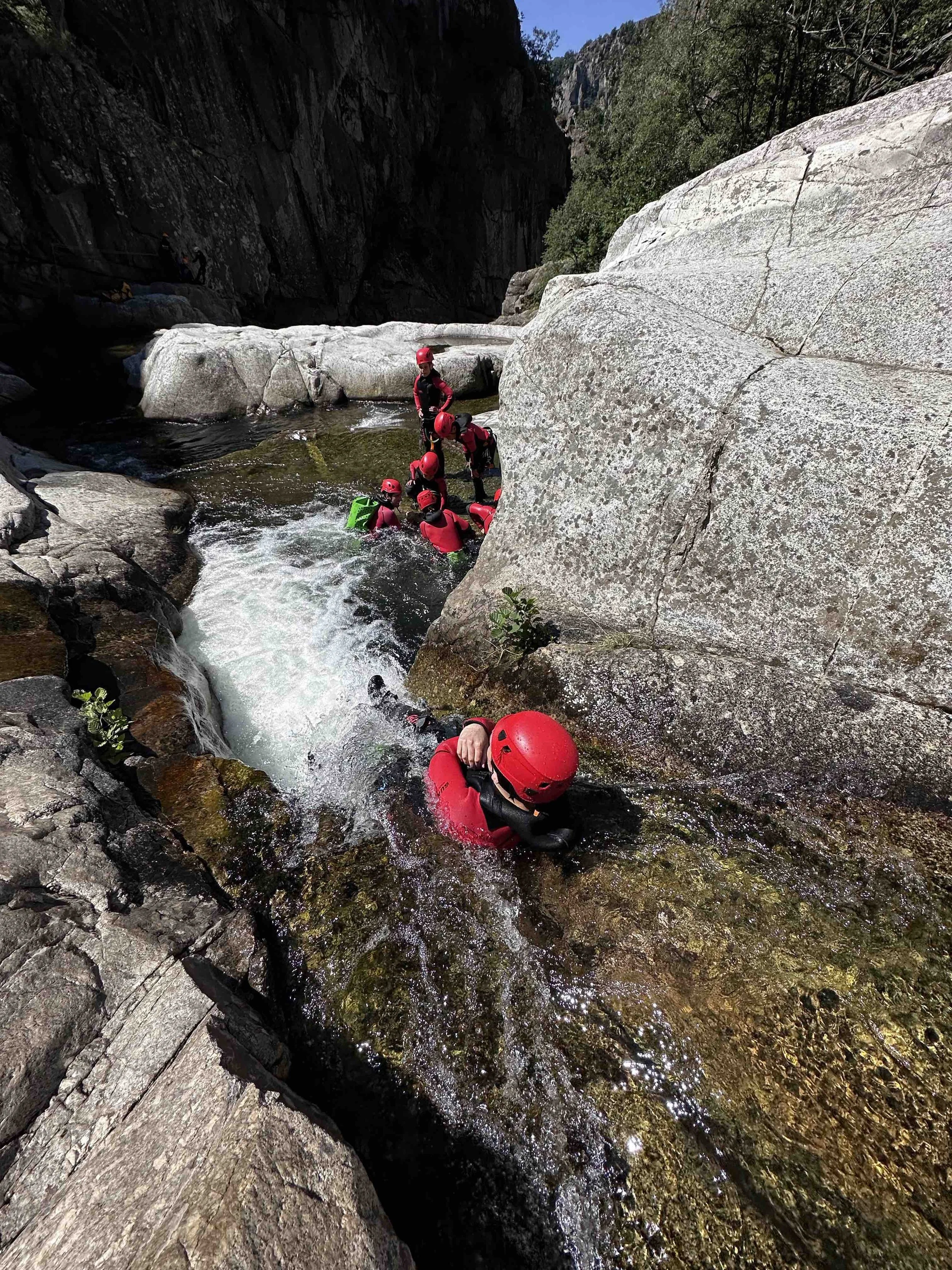Groupe de personnes pratiquant la canyoning dans une rivière entourée de rochers et de falaises, toutes équipées de casques et de combinaisons de plongée rouges.