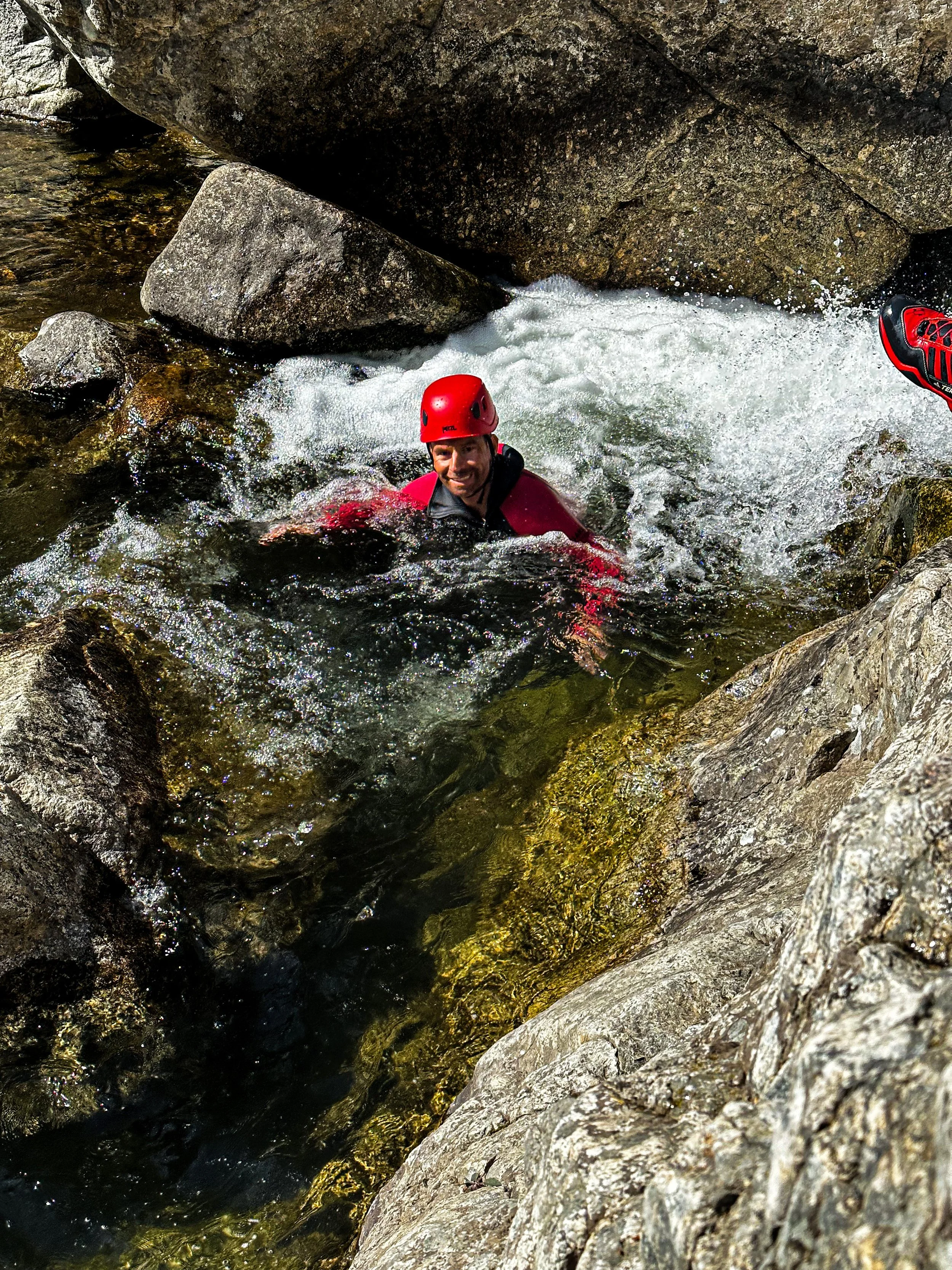 Un homme portant un casque rouge et une veste rouge nage dans une rivière entourée de rochers.
