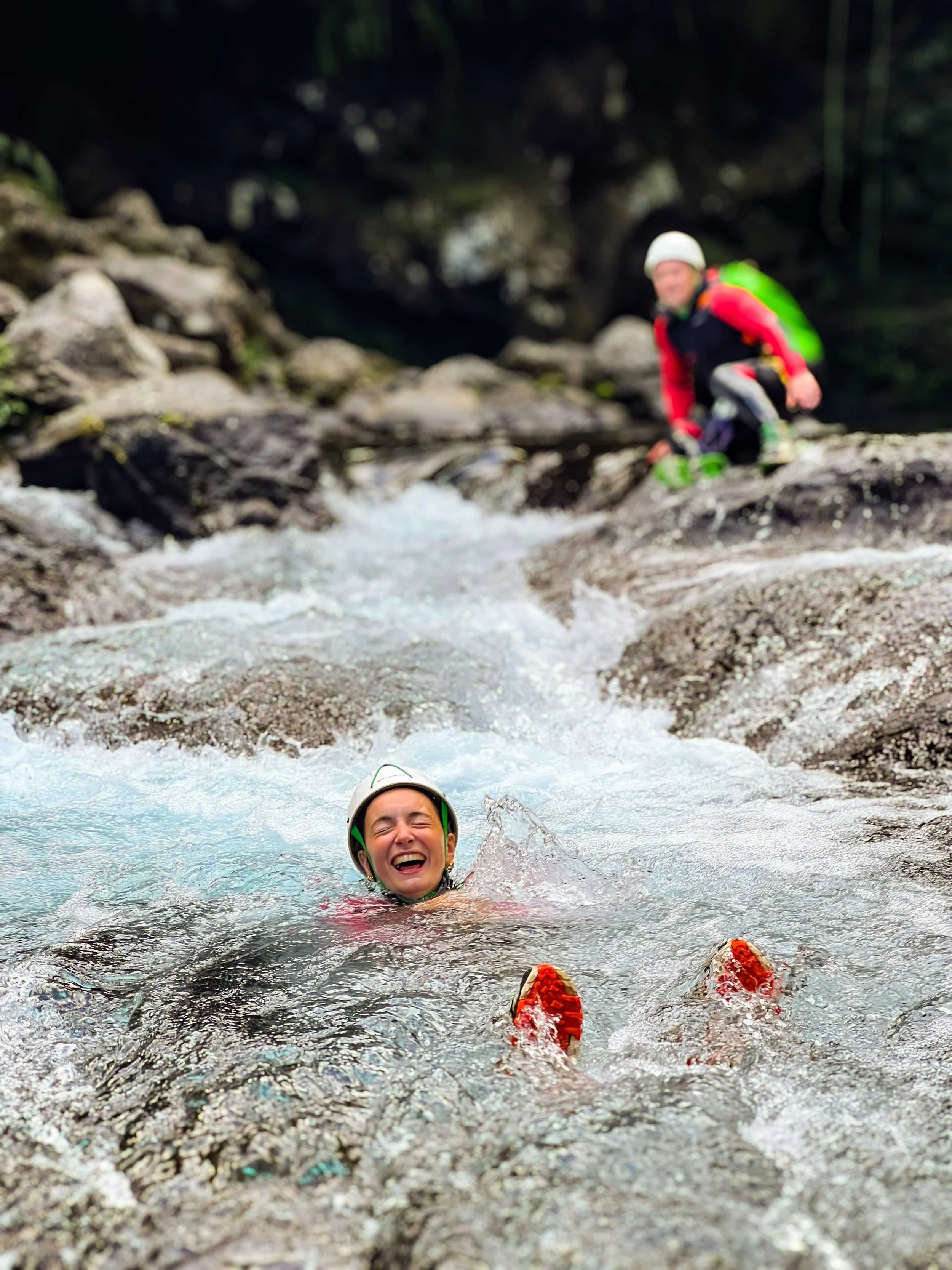 Une personne nageant dans une rivière en escaladant une roche, avec une autre personne en arrière-plan. Les deux portent des casques et des équipements de sécurité.