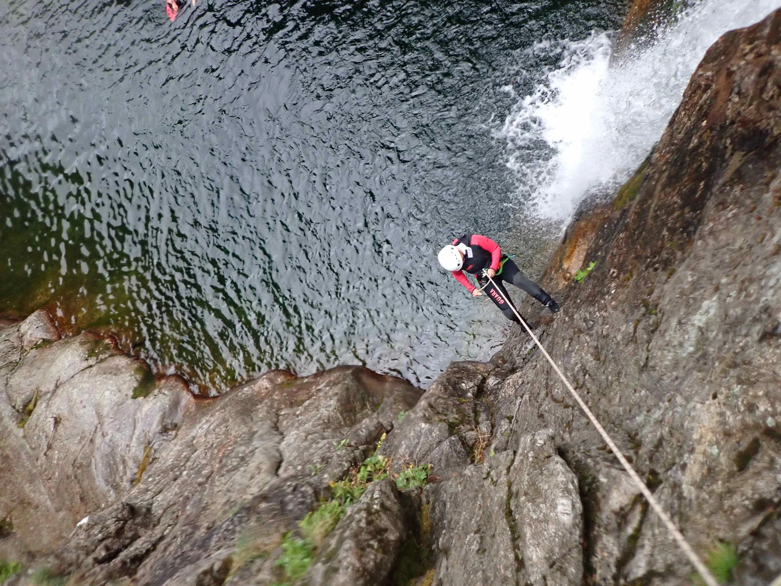 Une personne en équipement de canyoning, portant un casque blanc, descend une paroi rocheuse à côté d'une rivière ou d'une cascade.