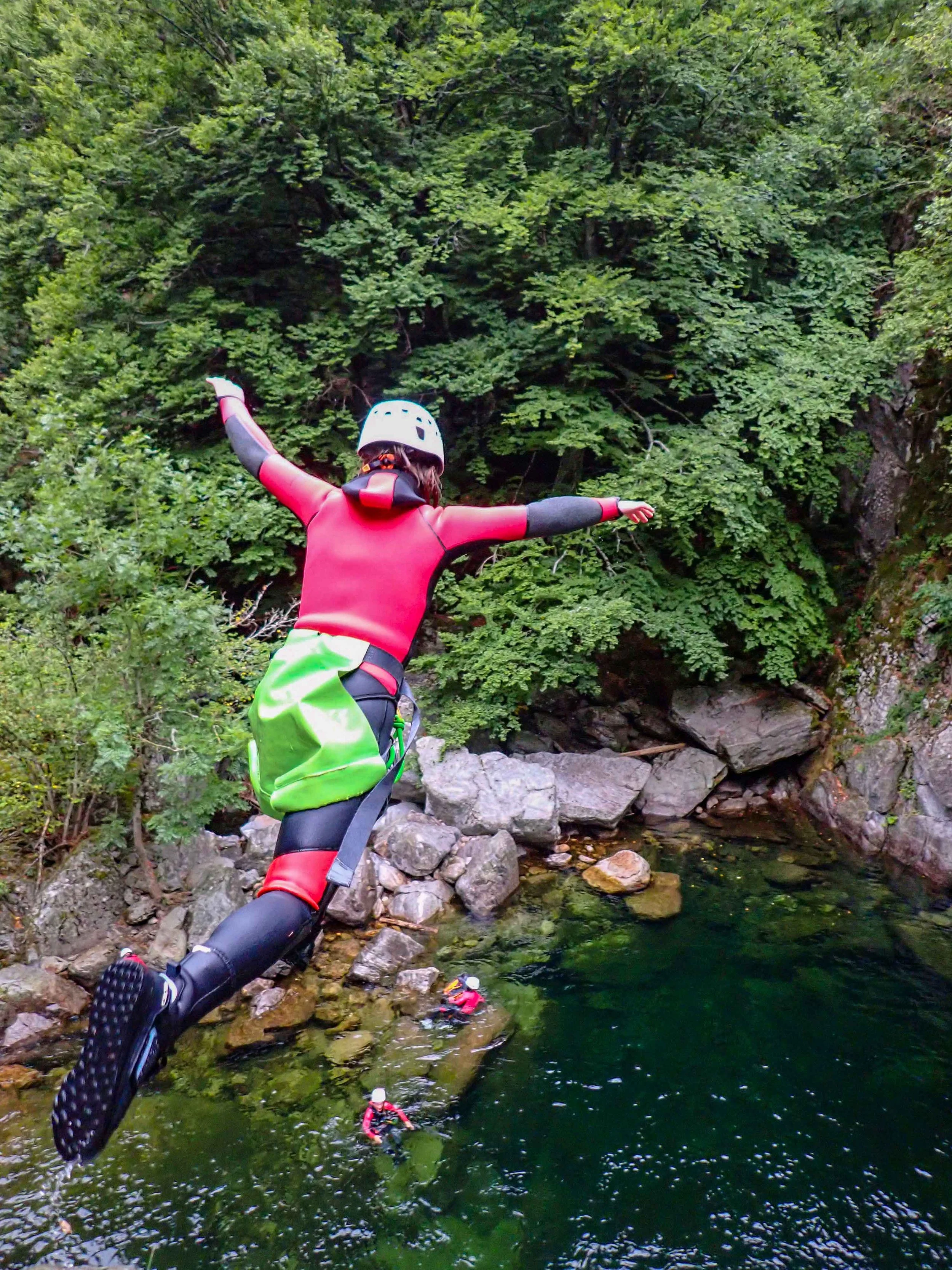 Une personne en équipement de canyoning sautant d'une roche dans une rivière entourée de végétation verte.