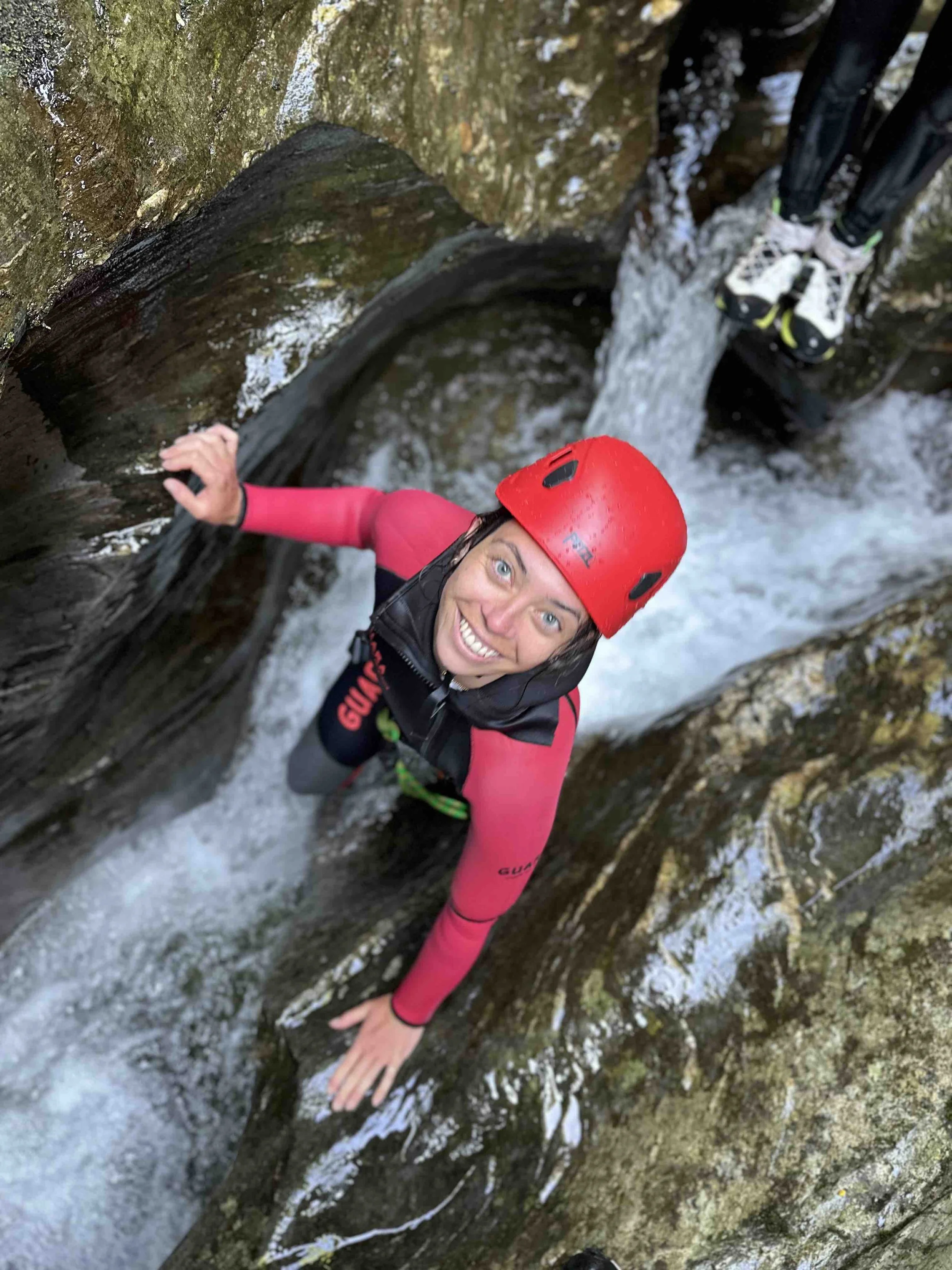 Une femme portant un casque rouge, une veste noire et une tenue de sport rose grimpe sur une roche mouillée dans une gorge étroite avec de l'eau qui coule.