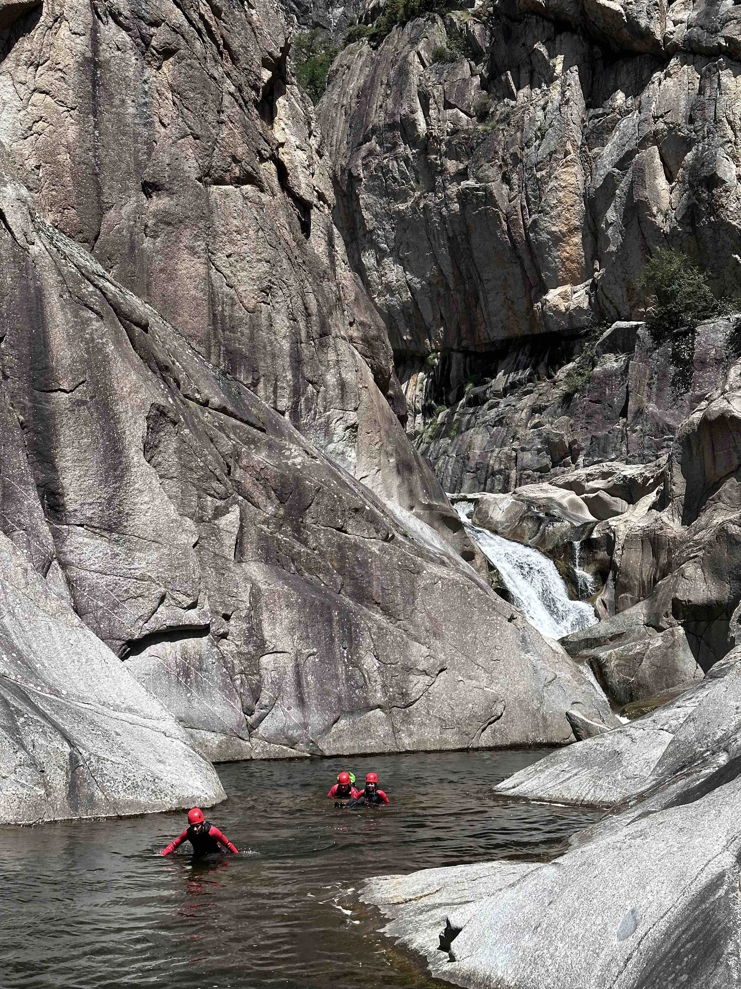Trois personnes en combinaison de canyoning rouge et casque rouge dans une rivière au fond d'une gorge rocheuse avec une petite cascade à l'arrière.