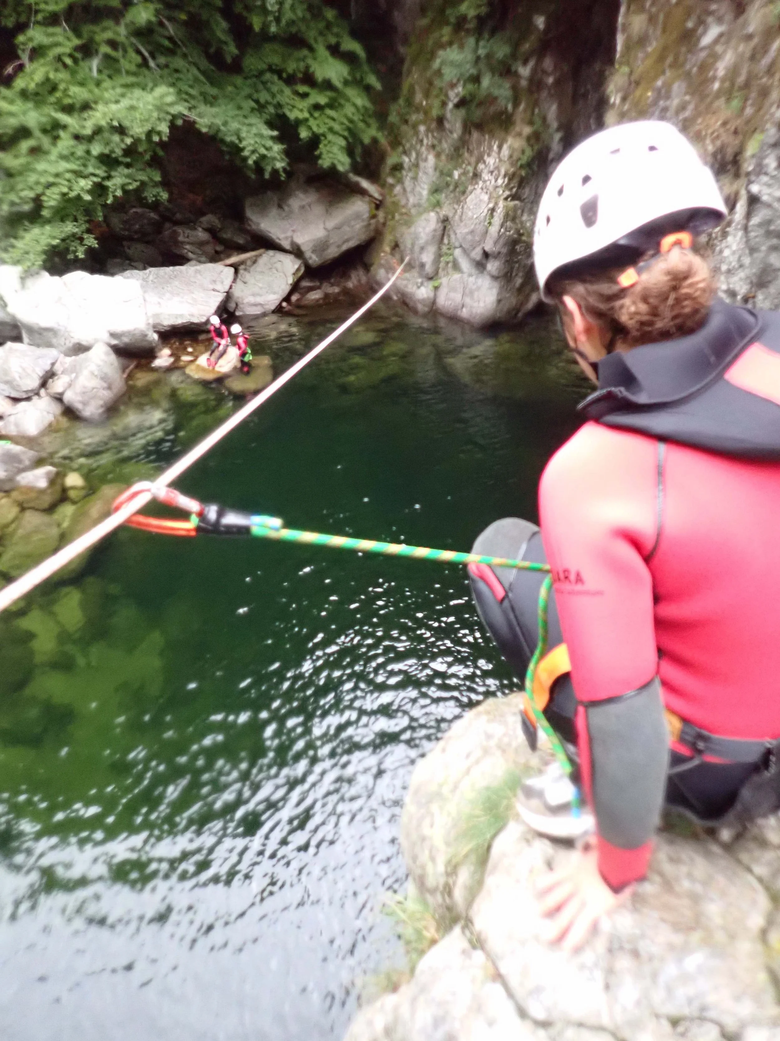 Personne portant un casque de sécurité, en train de pêcher ou d'explorer près d'une rivière ou d'un lac entouré de rochers et de végétation.