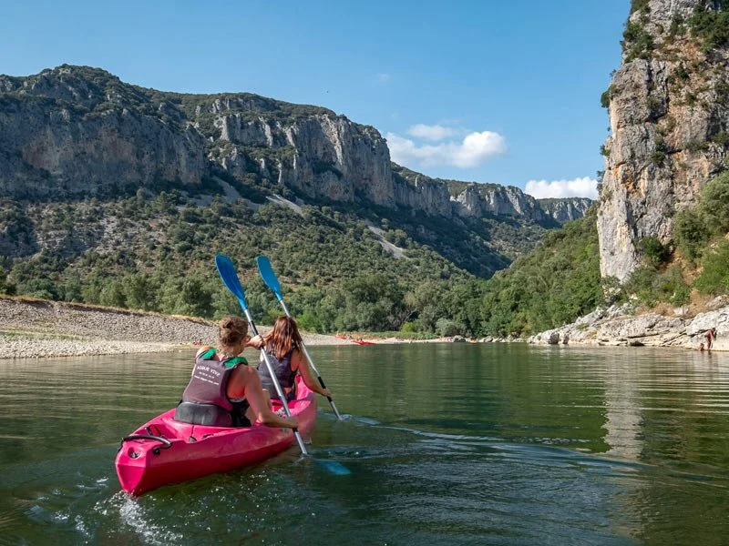 Deux personnes pagayant dans un kayak rose sur une rivière entourée de falaises rocheuses et de verdure.