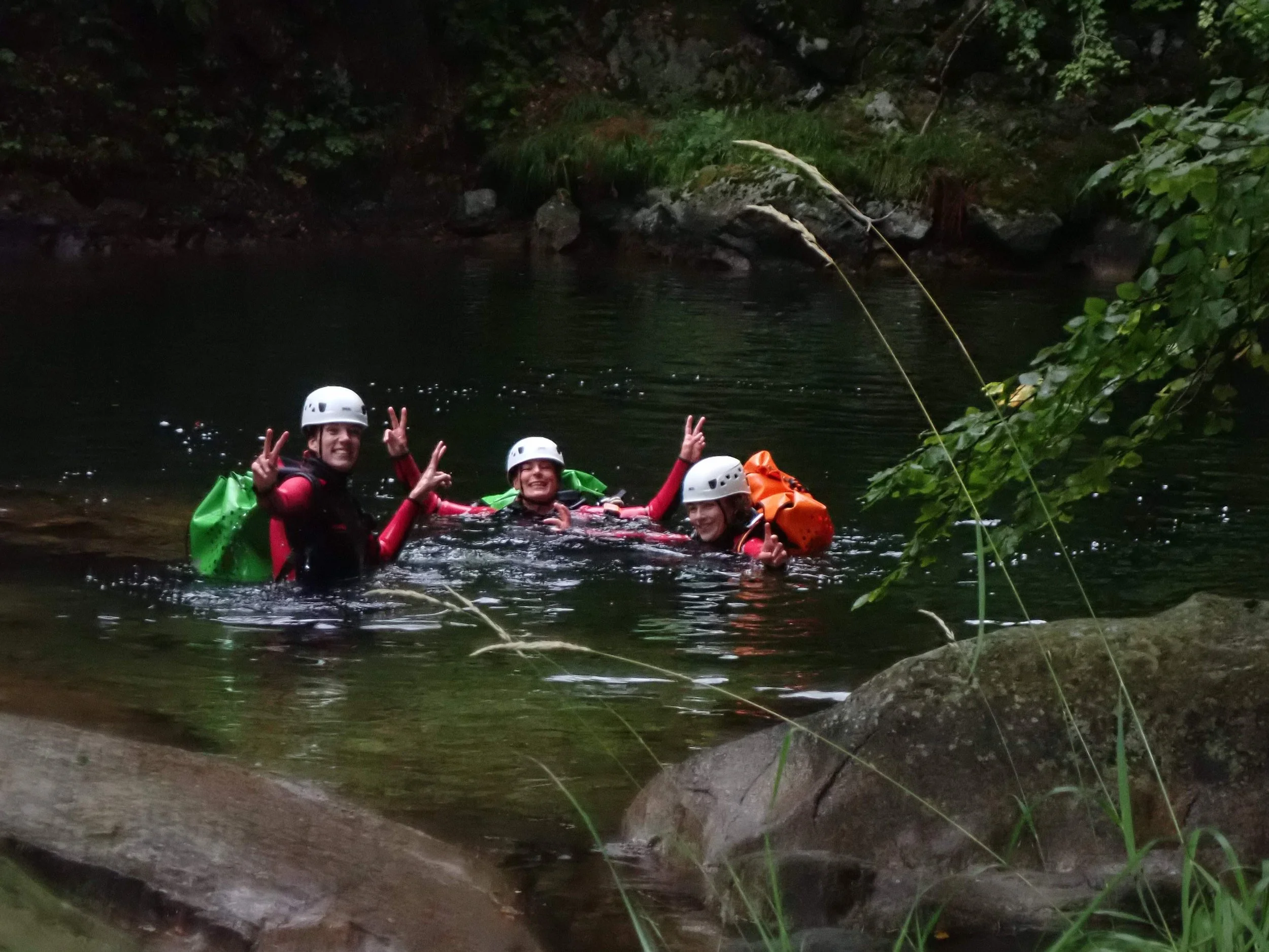 Trois personnes en équipement de sécurité flottant dans une rivière forestière, faisant un signe de paix avec leurs mains.