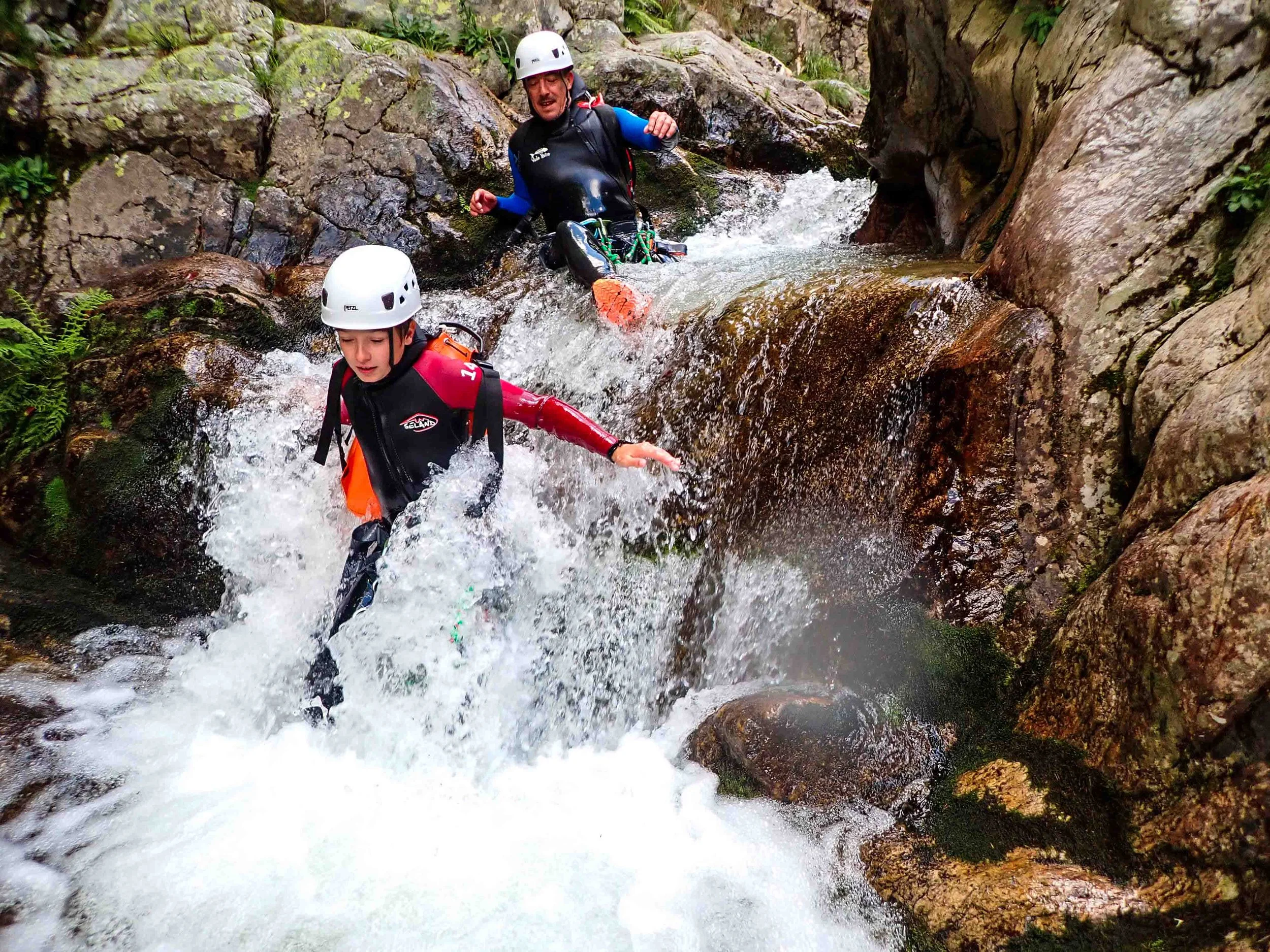 Deux personnes en équipement de sécurité descendent une rivière à l'aide de bottes et de casques, traversant des rochers et des eaux rapides.