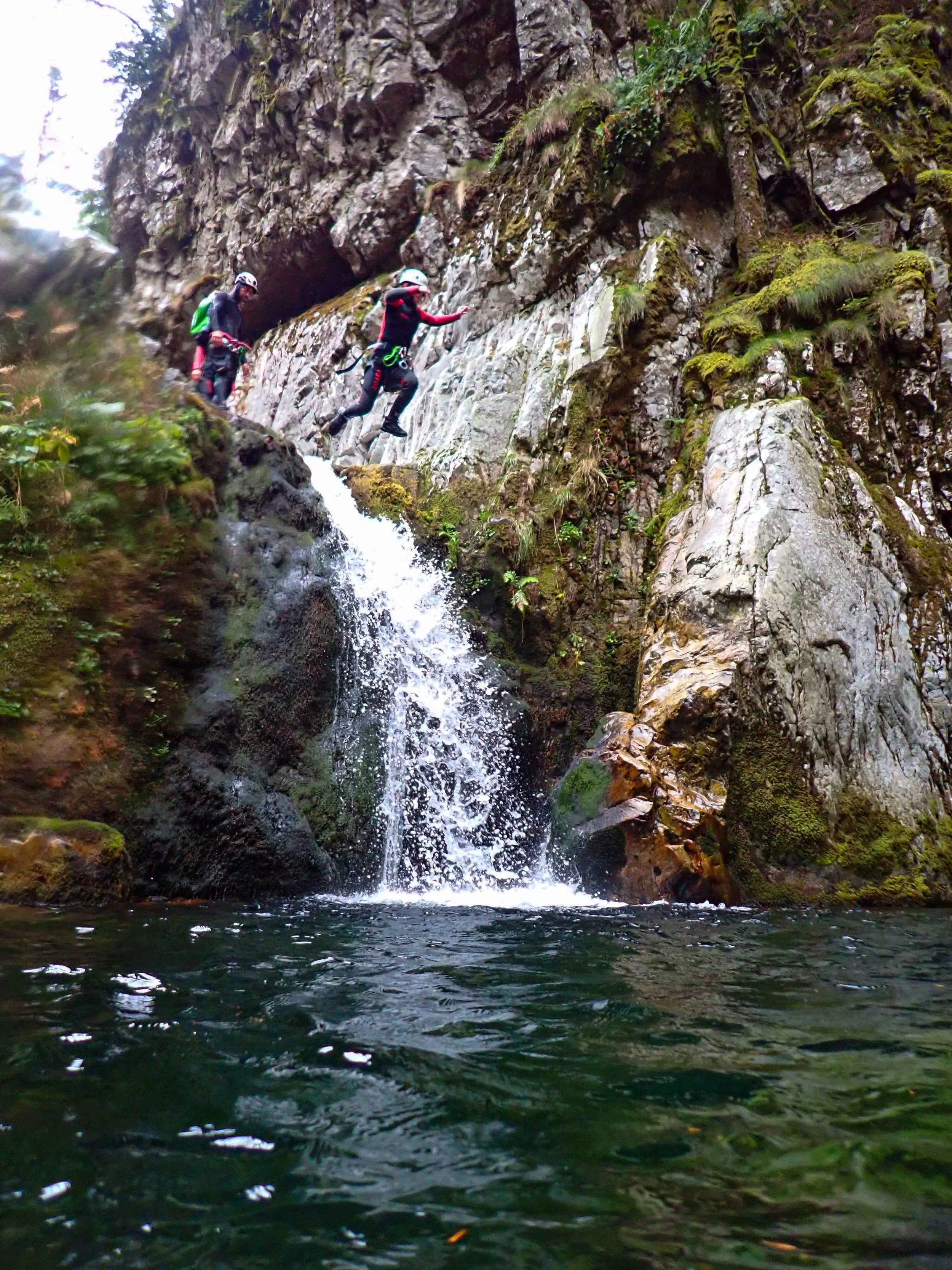 Deux personnes faisant de la descente en canyon dans une zone rocheuse avec une cascade et un étang en contrebas.