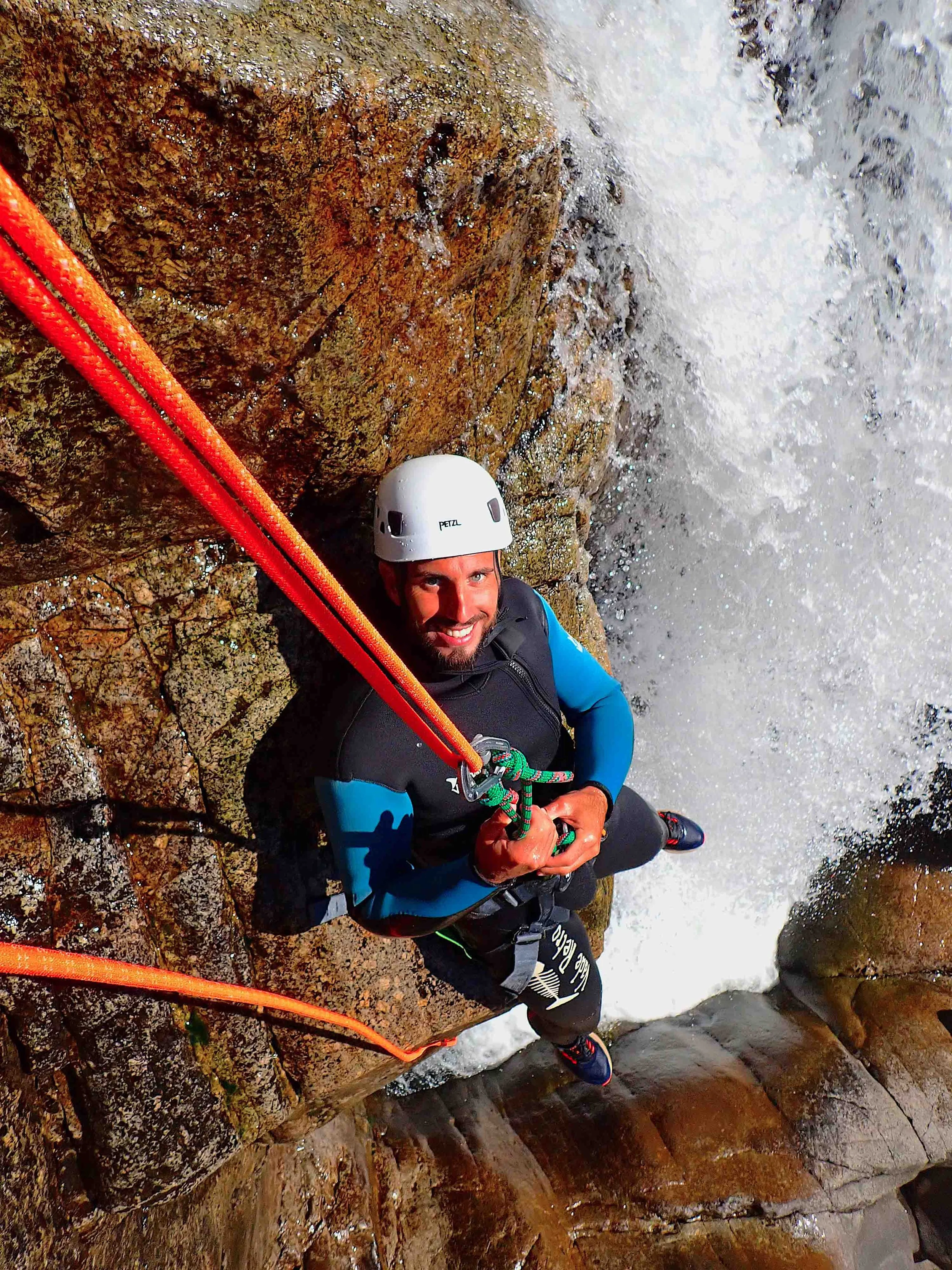 Un homme faisant de la cascades ou de l'escalade dans un canyon. Il porte un casque blanc, un harnais et une veste de protection, accroché à une corde orange.