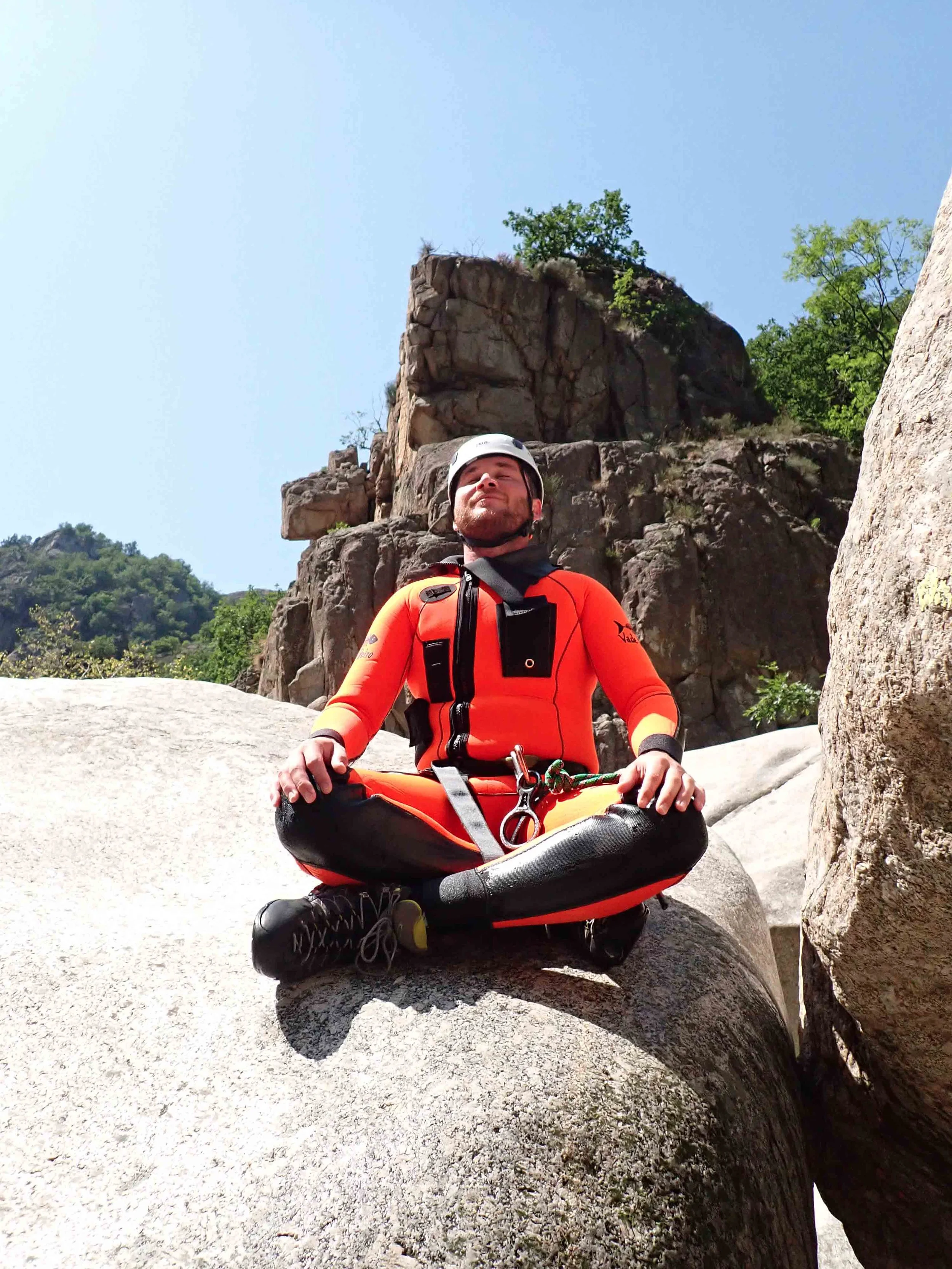 Un homme en tenue d'escalade orange et noir, portant un casque blanc, assis en méditation sur une roche dans un paysage de montagnes et de rochers, avec un ciel bleu clair en arrière-plan.