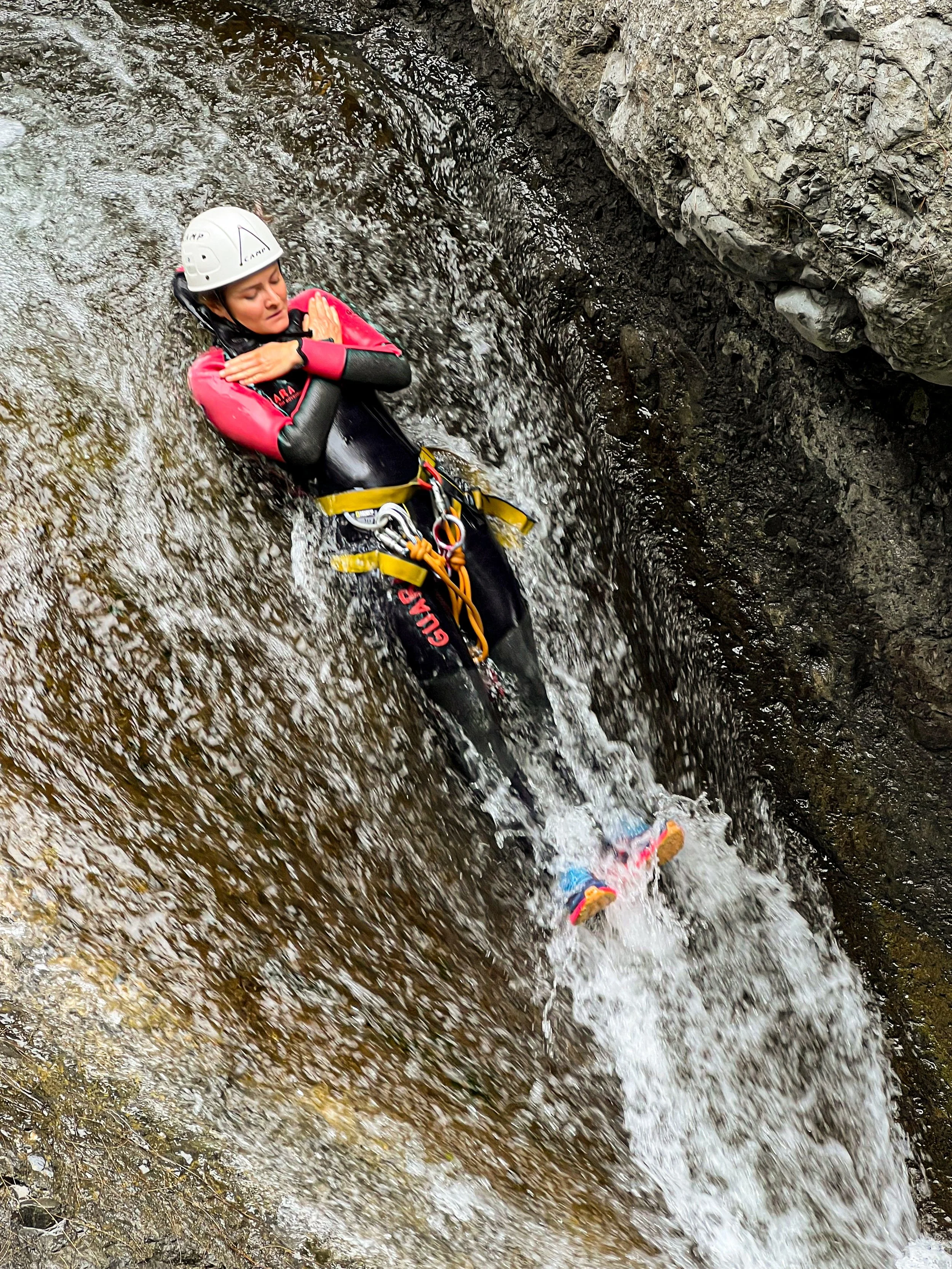 Une femme en équipement de canyoning, portant un casque blanc, un wetsuit noir et rose, et une ceinture de sécurité, se repose dans un ruisseau rocheux.