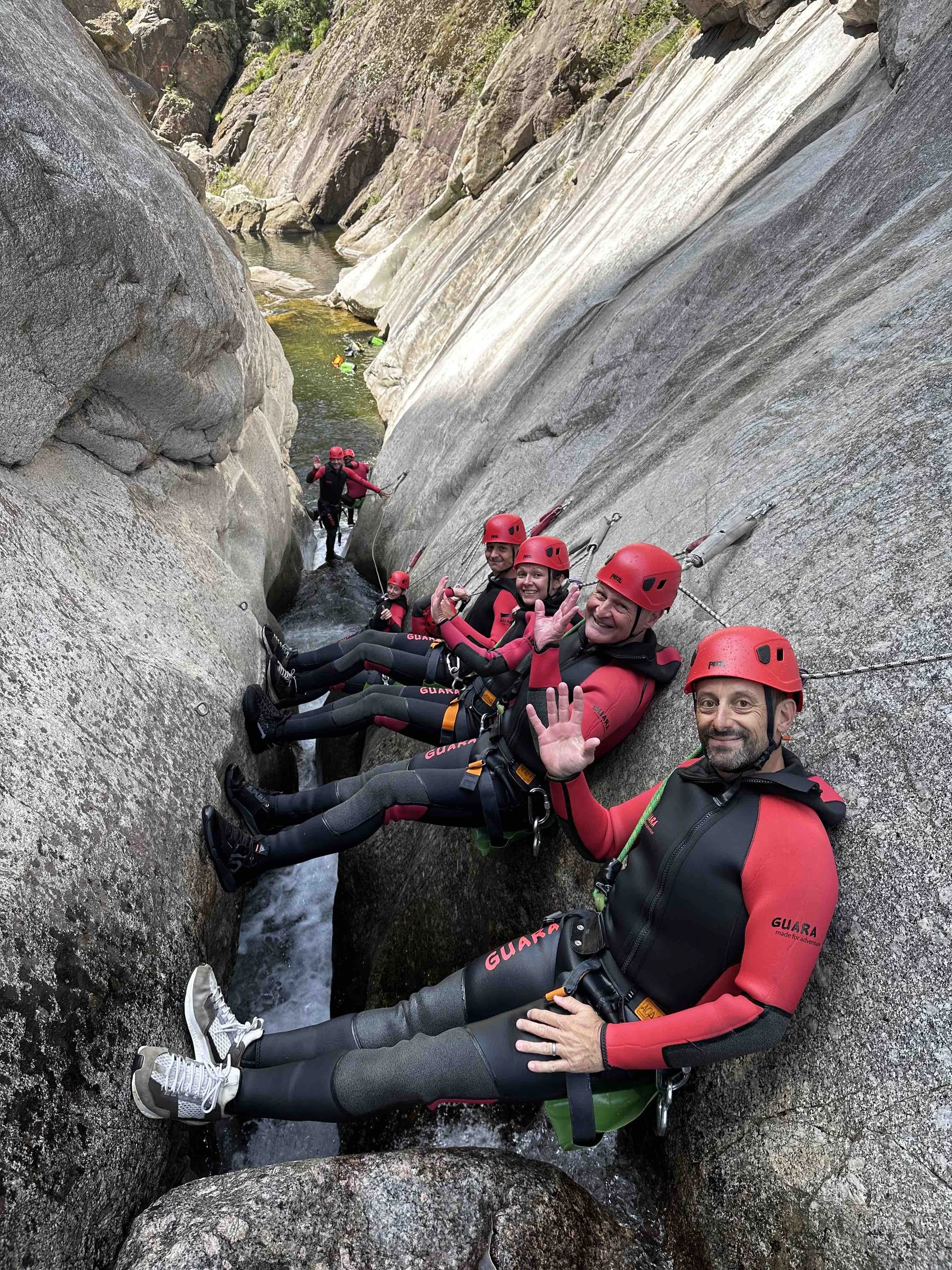 Groupe de personnes équipées pour le canyoning, assises dans une faille entre deux rochers, portant des casques rouges et des combinaisons noires et rouges, saluant la caméra. En arrière-plan, d'autres personnes explorent la rivière.
