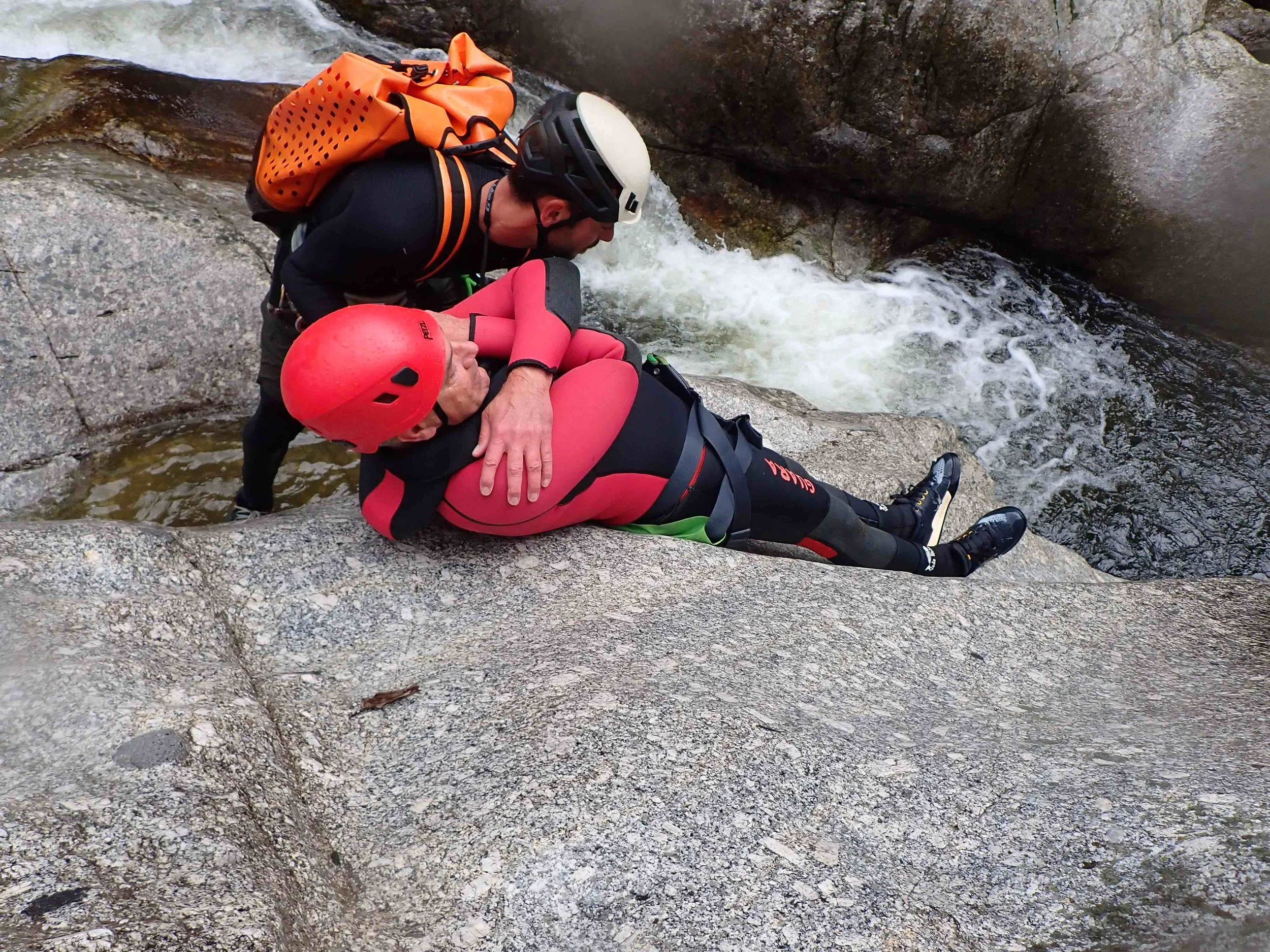 Un homme secourt une autre personne blessée lors d'une activité de canyonisme, près d'une rivière à cascade, tous deux portant un casque et des vêtements de sport.