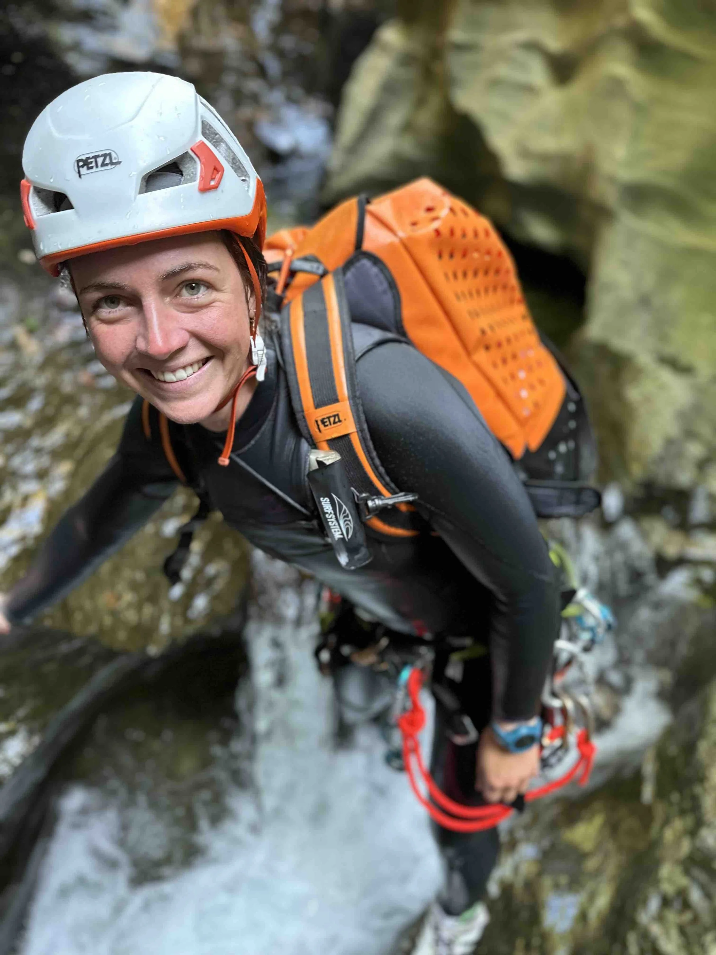 Une femme en équipement d'escalade, portant un casque blanc, un harnais et un sac à dos orange, grimpe sur une paroi rocheuse près d'une cascade.