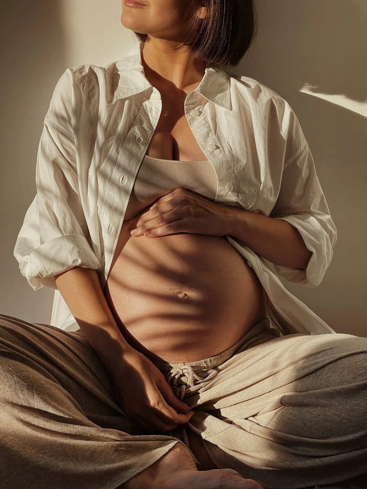 A woman with short hair, wearing a white shirt over a beige tank top and beige pants, sitting with her legs crossed, illuminated by sunlight casting shadows on her abdomen.