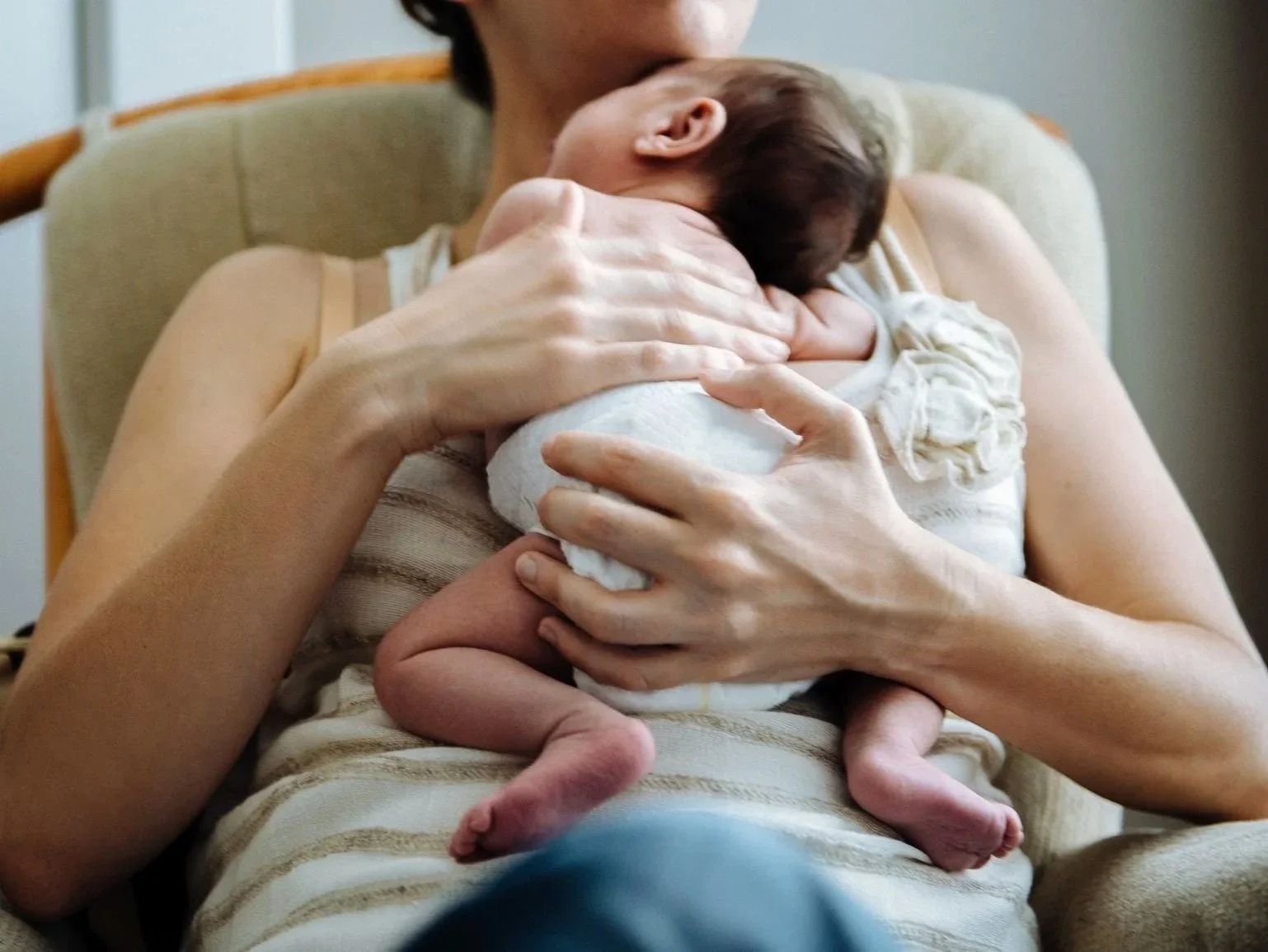 A woman holding a newborn baby while sitting on a beige armchair in a softly lit room.