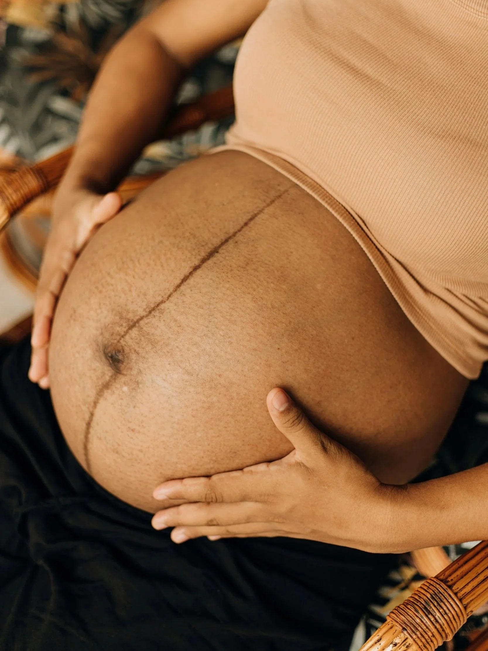 Close-up of a pregnant person's belly with a black line drawn from the navel to the top of the belly, resting on a wicker chair. The person is wearing a beige top and black pants.