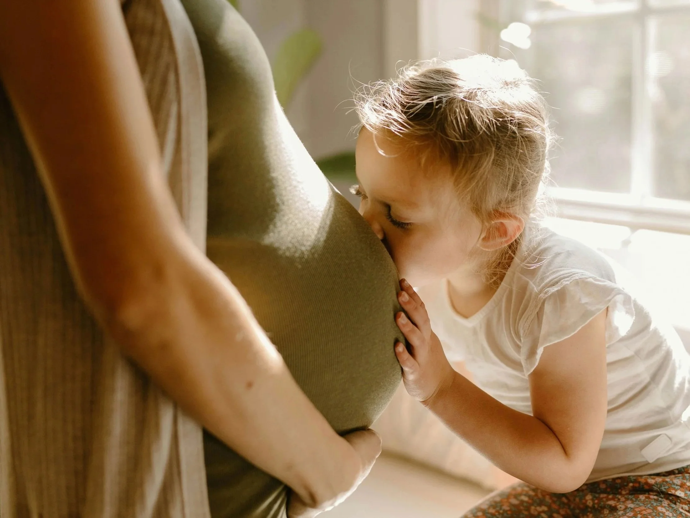 A young girl kissing a pregnant woman's belly in a sunlit room.