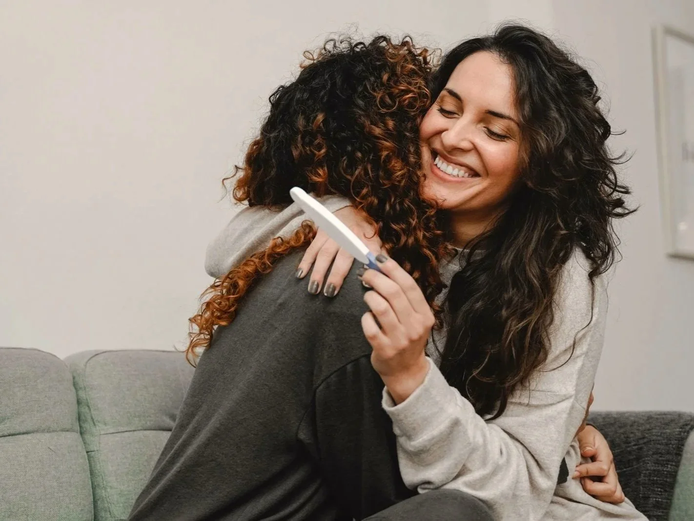 Two lesbian women hugging, one holding a positive pregnancy test, smiling with joy in a cozy living room.