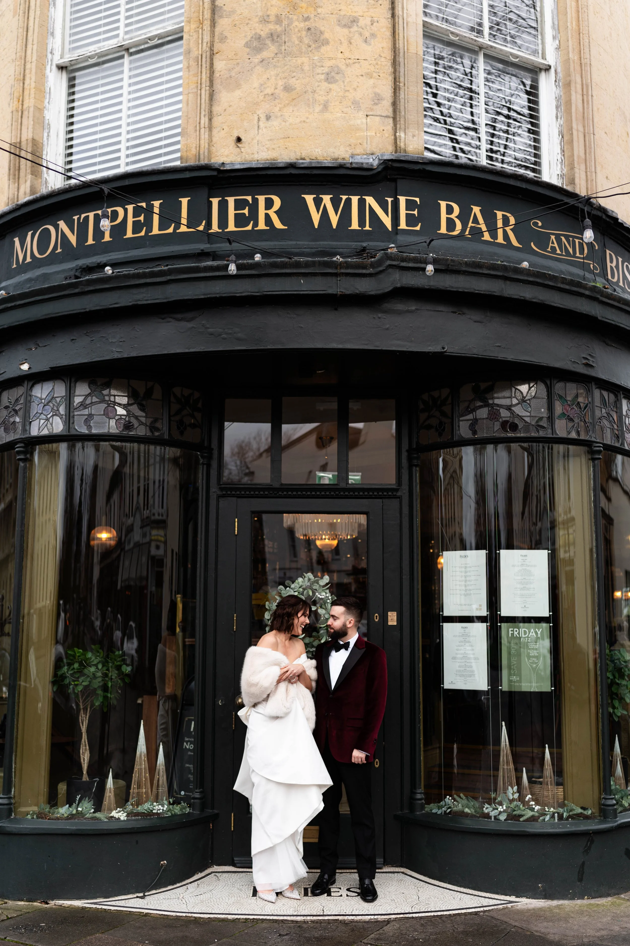 A newlywed couple dressed in wedding attire stands outside a black storefront of Montpelllier Wine Bar and Bistro, sharing a moment of affection.