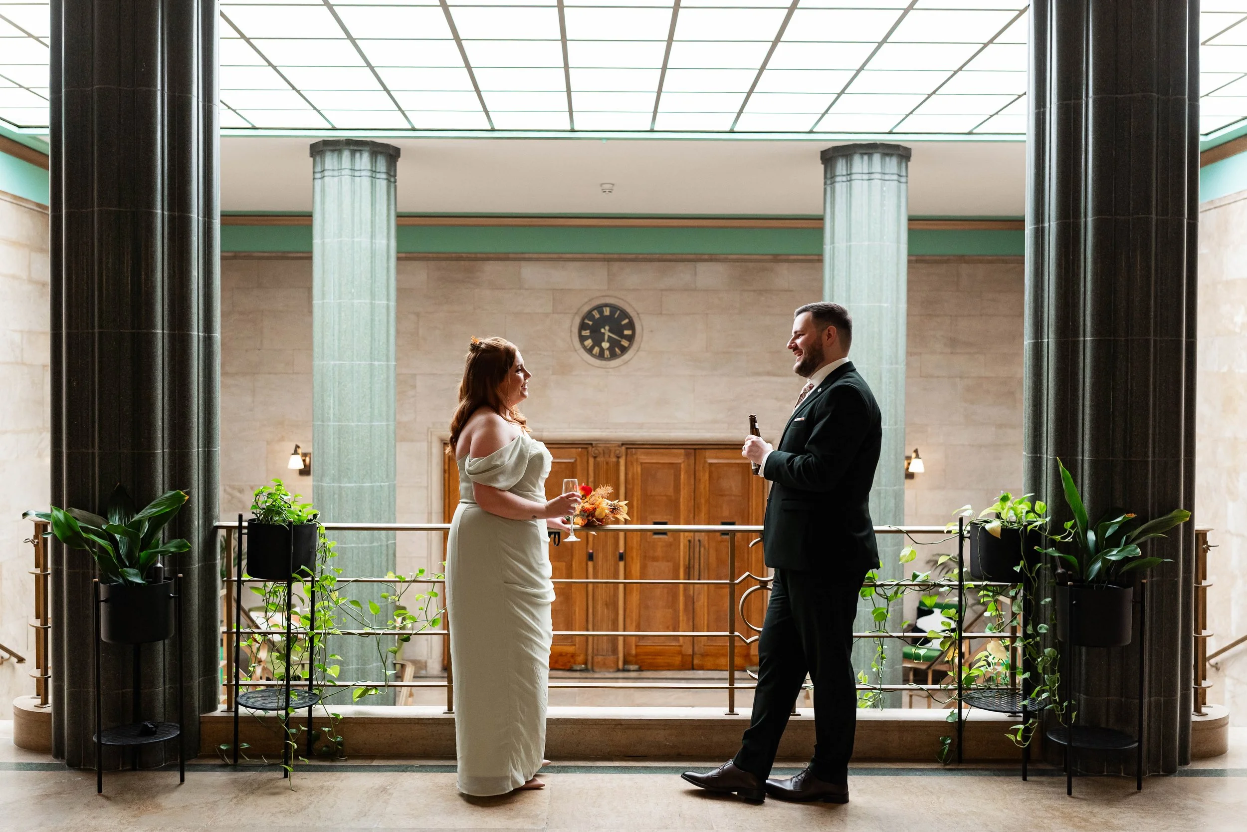 A couple in formal attire standing face to face in an elegant interior with large green columns, holding drinks, celebrating a special occasion.