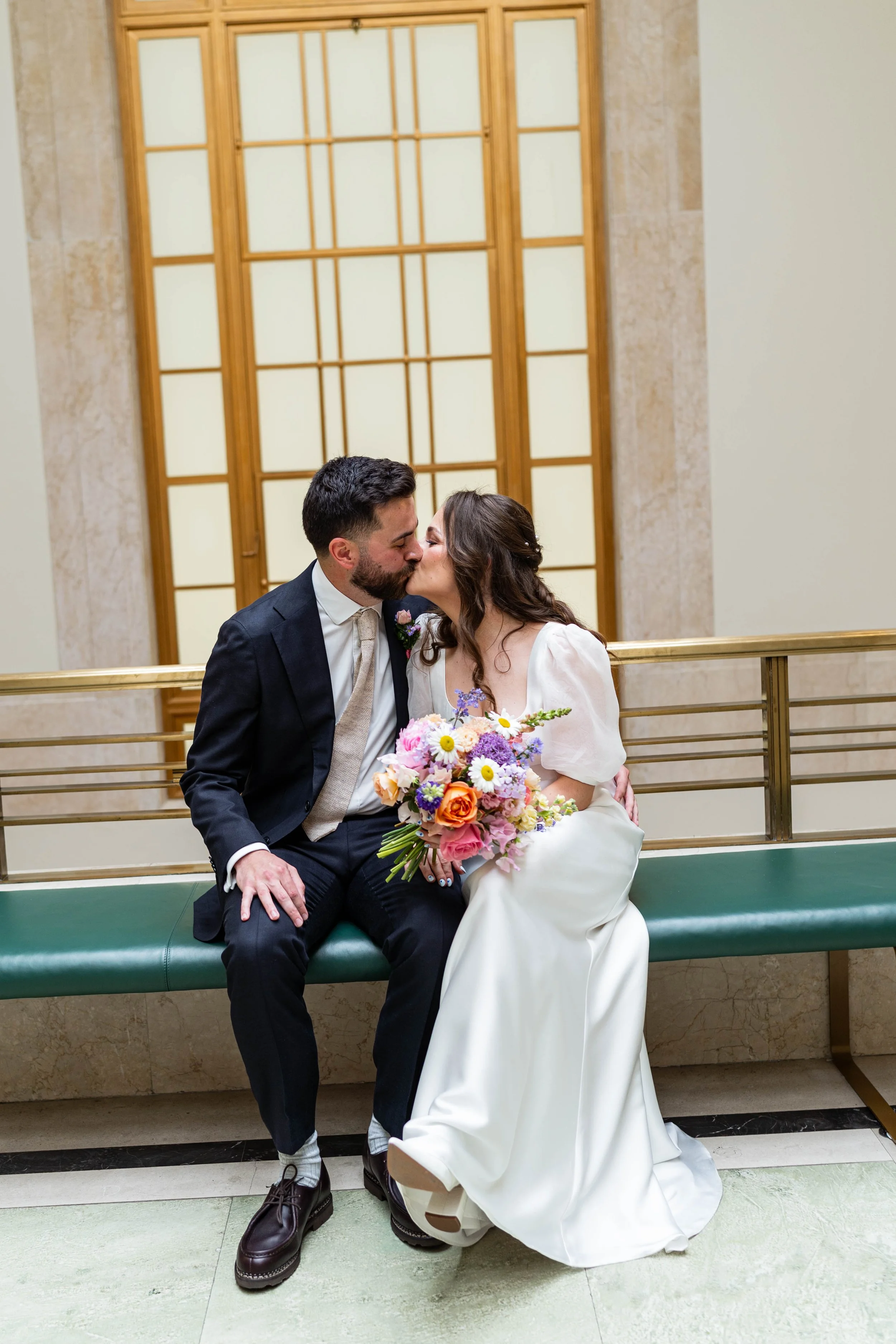 A newlywed couple sharing a kiss on a bench indoors. The bride is holding a bouquet of colorful flowers. The groom is wearing a dark suit with a patterned tie. The bride is wearing a white wedding dress with puff sleeves.