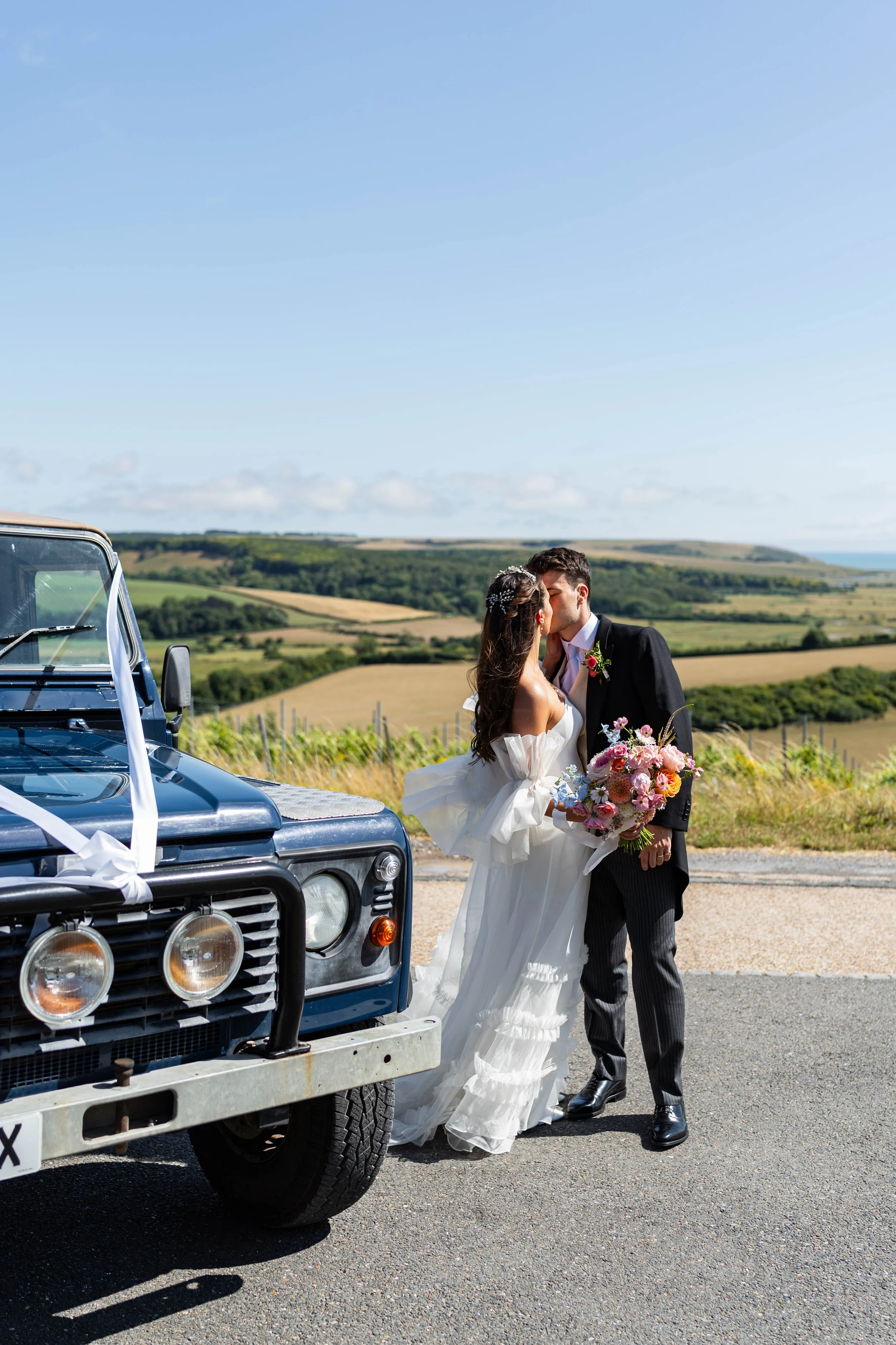A bride and groom sharing a kiss next to a decorated vintage Land Rover vehicle on a rural countryside road with green fields and rolling hills in the background.