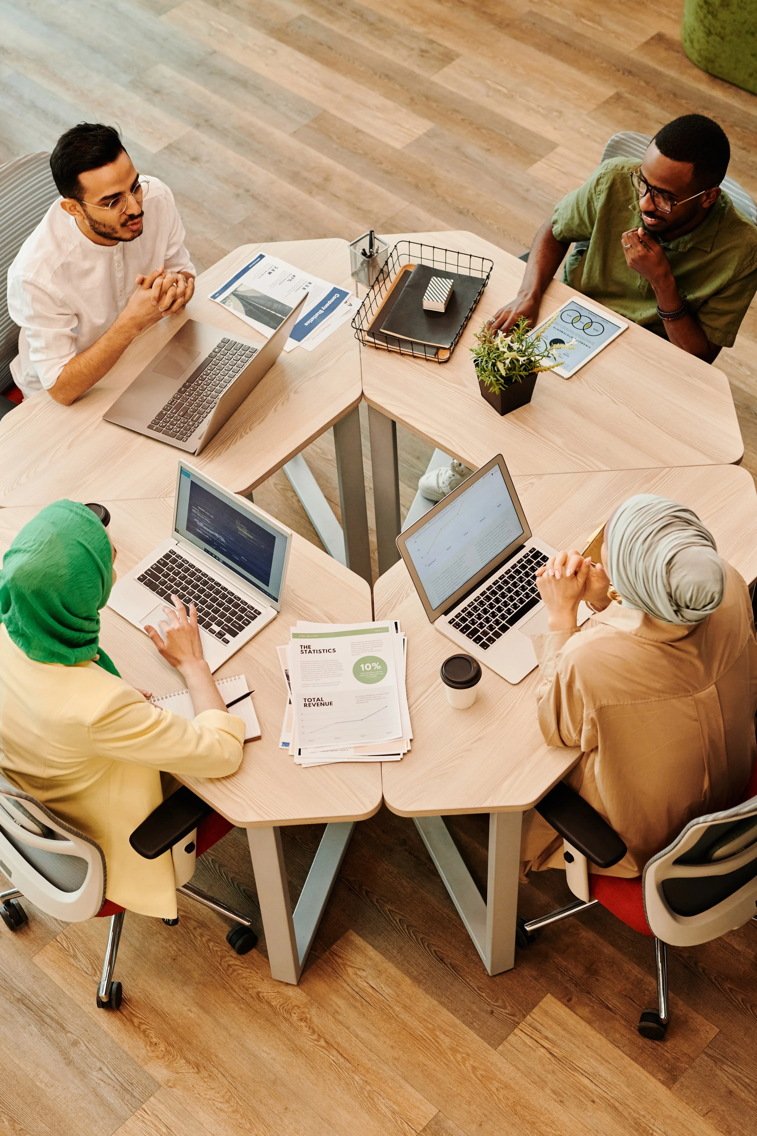 Five diverse professionals sit around a curved wooden conference table in a meeting, using laptops and discussing documents, with a few potted plants and office supplies on the table.