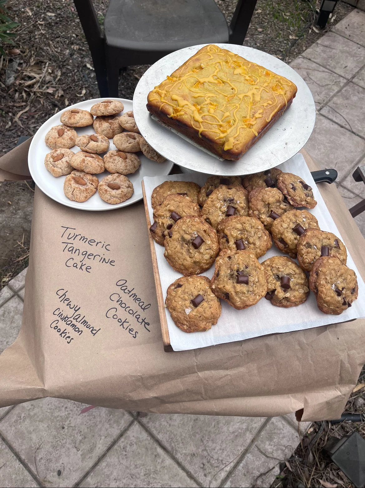 An outdoor table with a brown paper covering displaying various baked goods labeled as turmeric tangerine cake, oatmeal chocolate chip cookies, and chewy cinnamon cookies. The table holds a plate of oatmeal cookies, a tray of chewy cinnamon cookies, and a rectangular turmeric tangerine cake with yellow icing drizzle.
