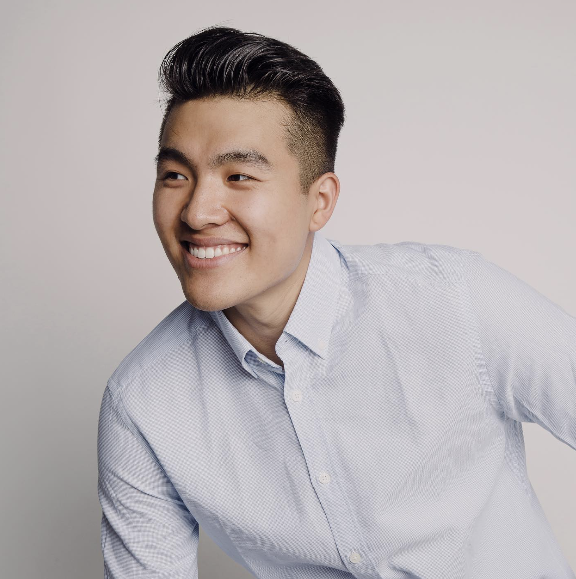 Smiling young Asian man wearing a light blue button-up shirt posing against a plain background.